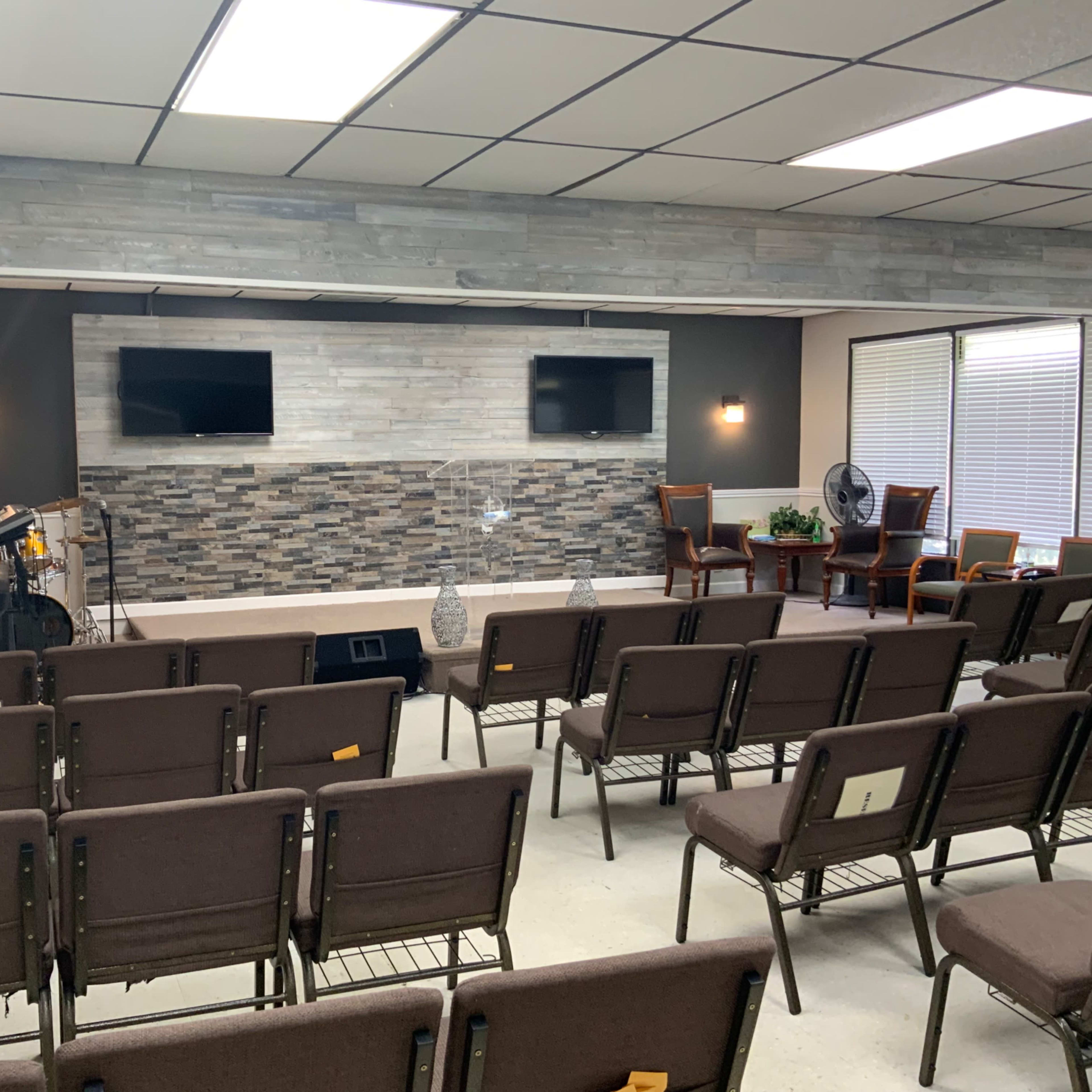 An interior room set up for a gathering, featuring rows of brown chairs, two side tables, and a stone accent wall with two mounted screens.