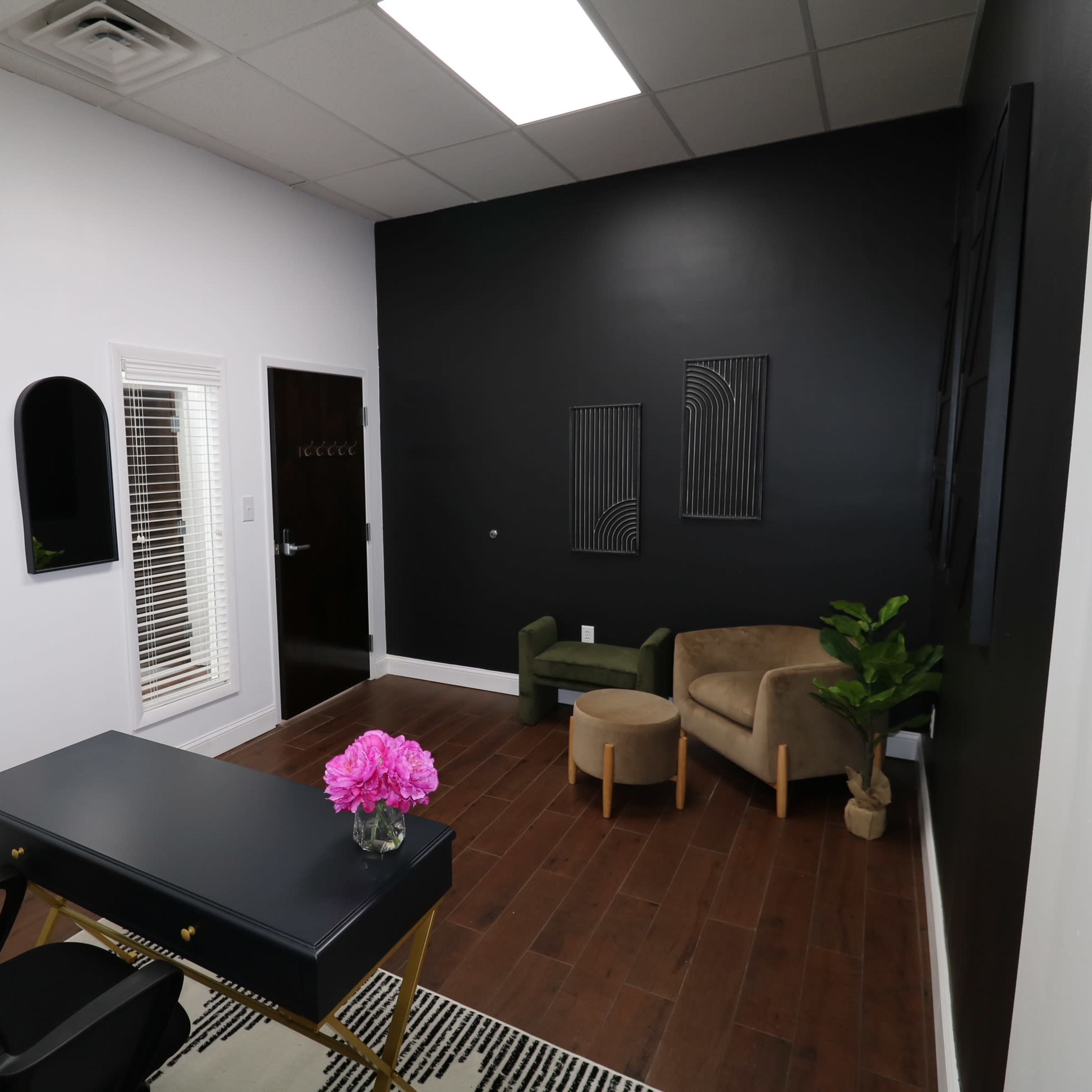 A modern office space with a black accent wall, a wooden floor, a black desk with a flower vase, and a small seating area with two chairs.