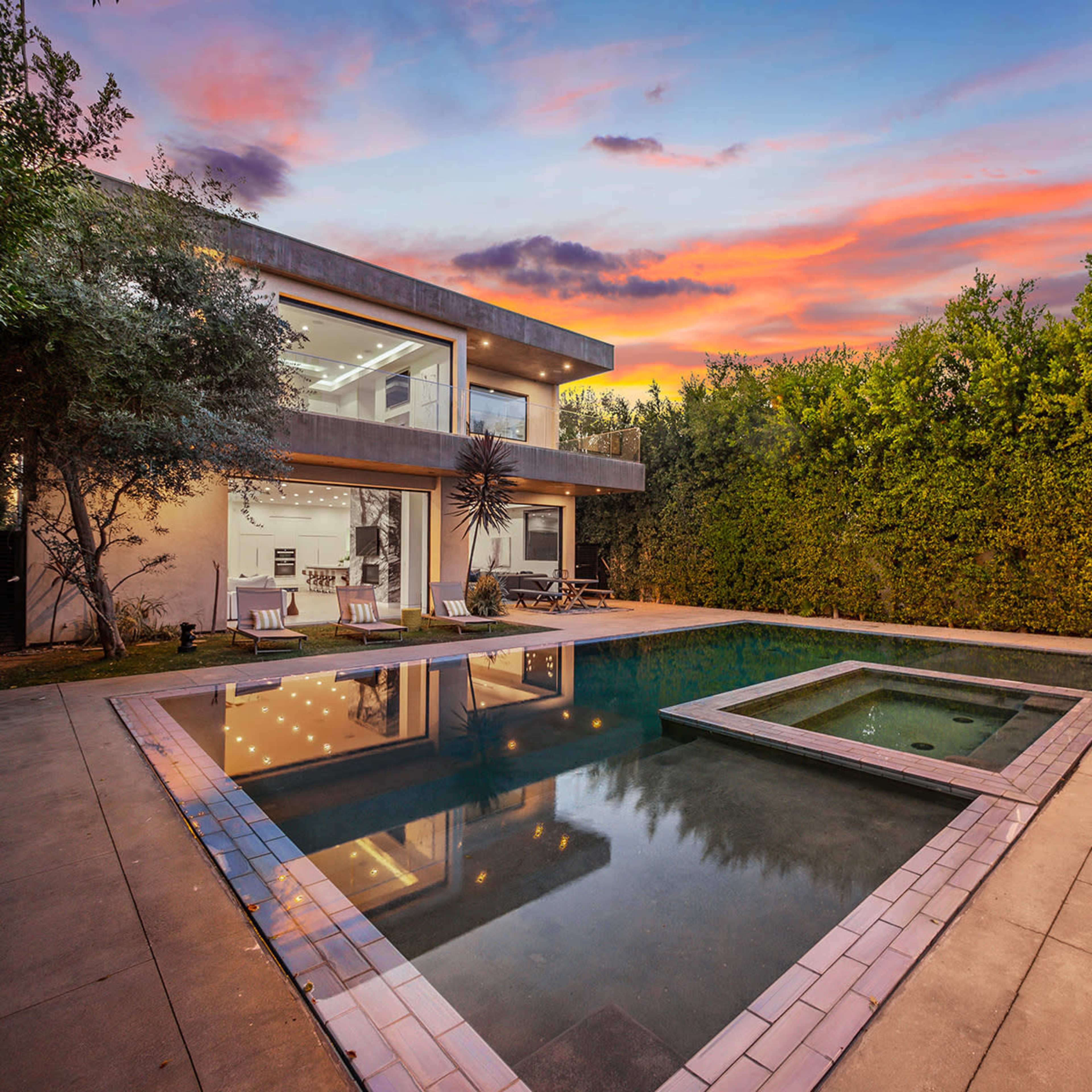 A modern house with large windows, a swimming pool, and a spa area is surrounded by greenery at sunset.