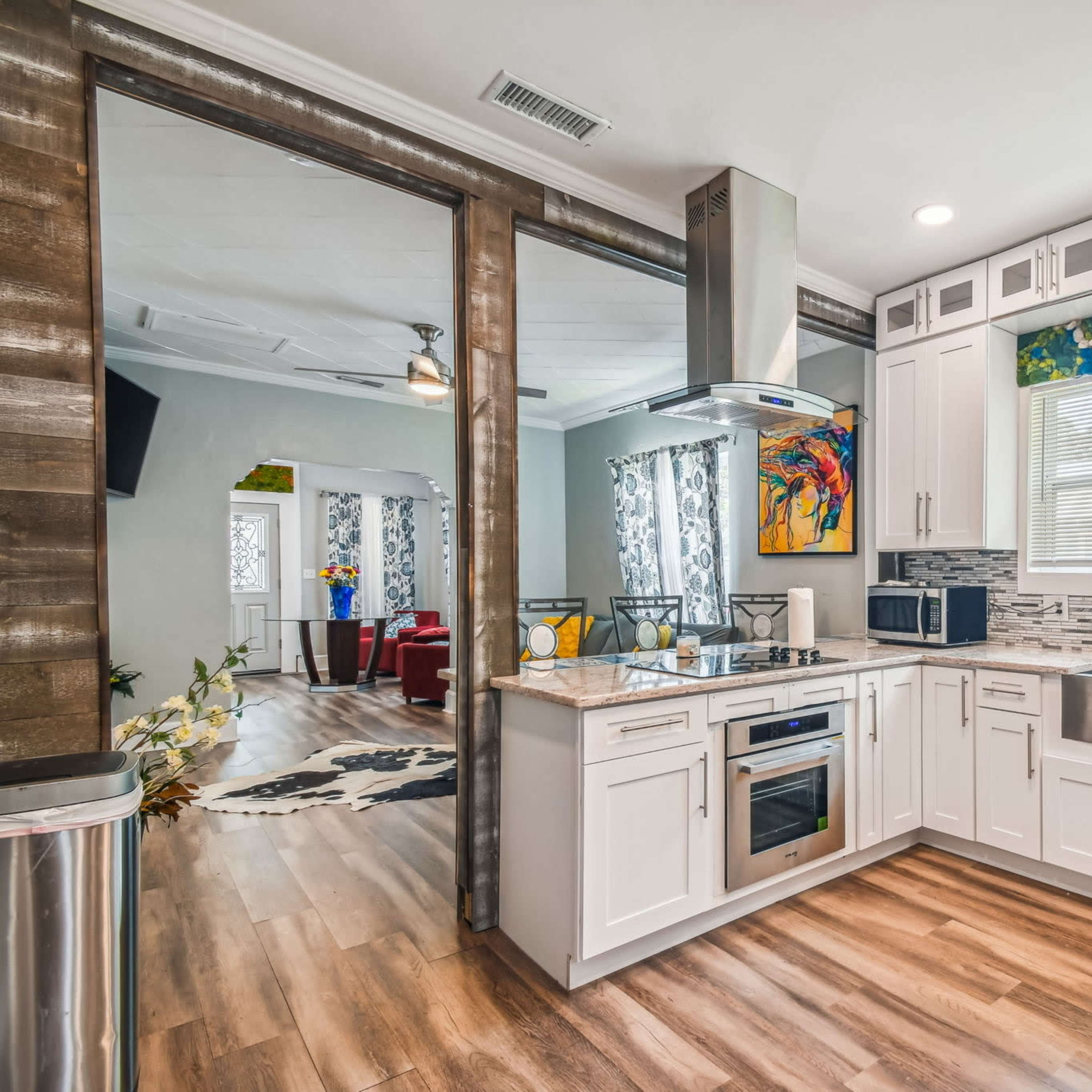 A modern kitchen features white cabinetry, stainless steel appliances, and a countertop with a large sink, adjacent to a living area with colorful decor.