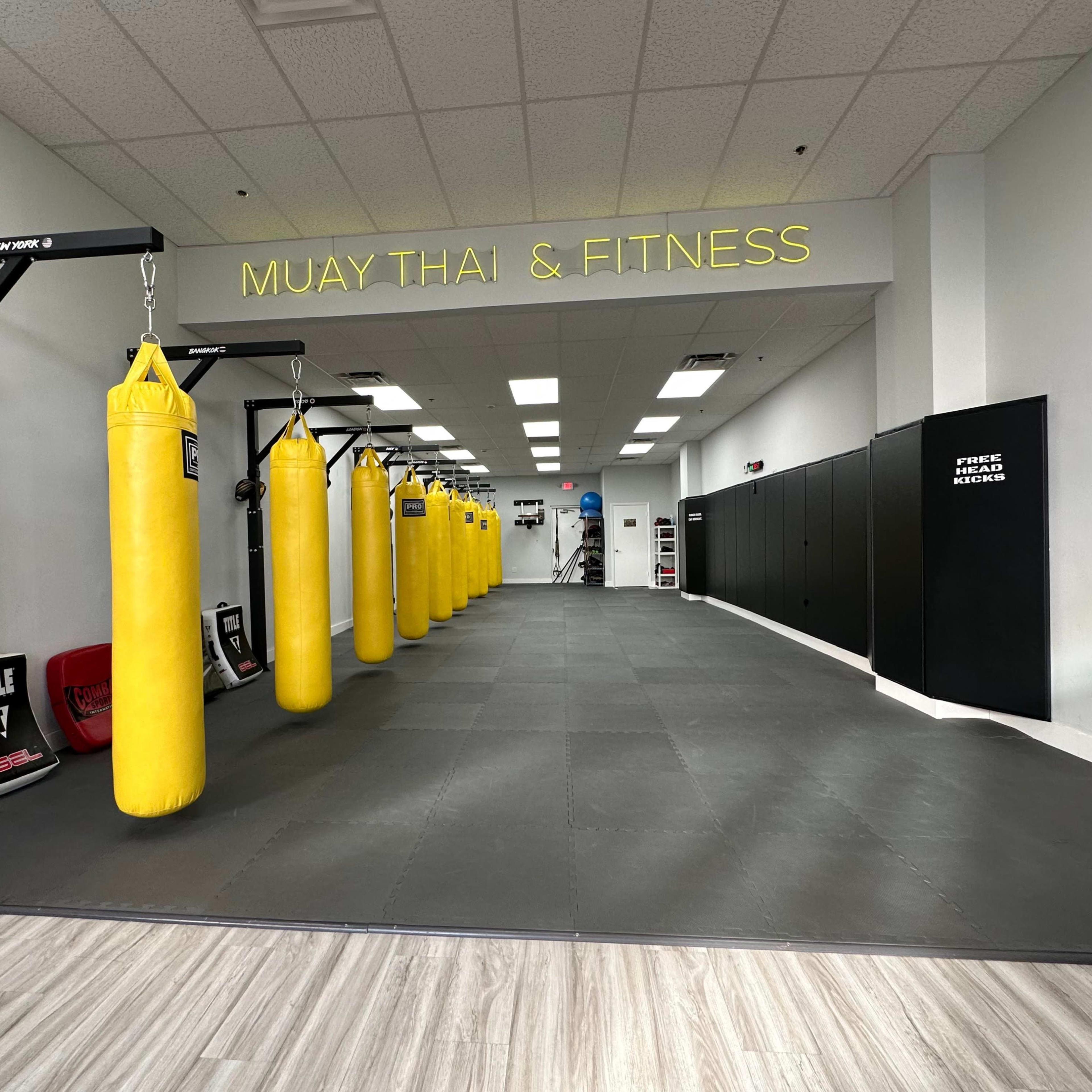A Muay Thai and fitness gym featuring a row of yellow punching bags along one side of a spacious training area with black mats and padded walls.