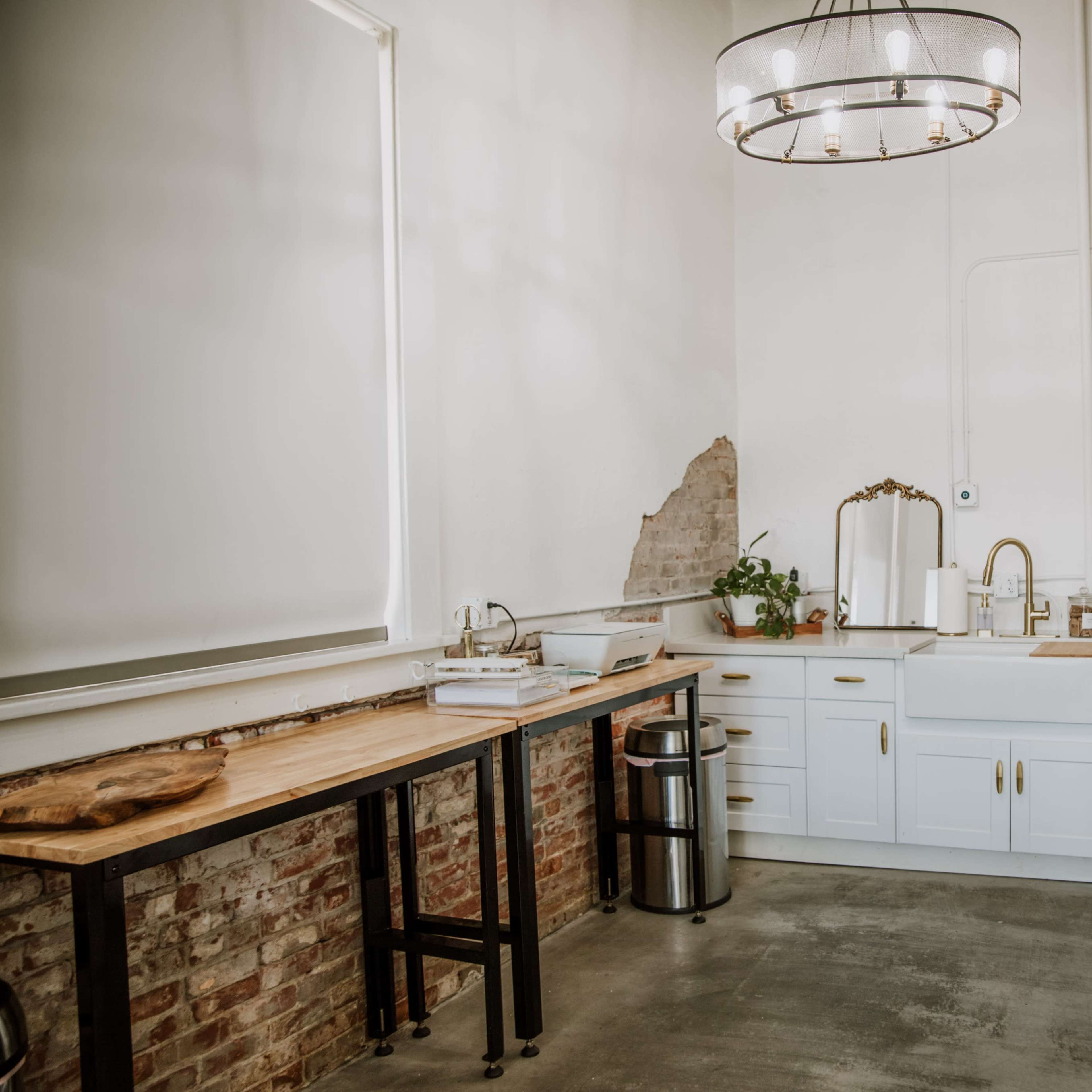 A bright studio kitchen area with a wooden table, modern cabinetry, and a refrigerator featuring the text "CASA BLOOM STUDIOS."