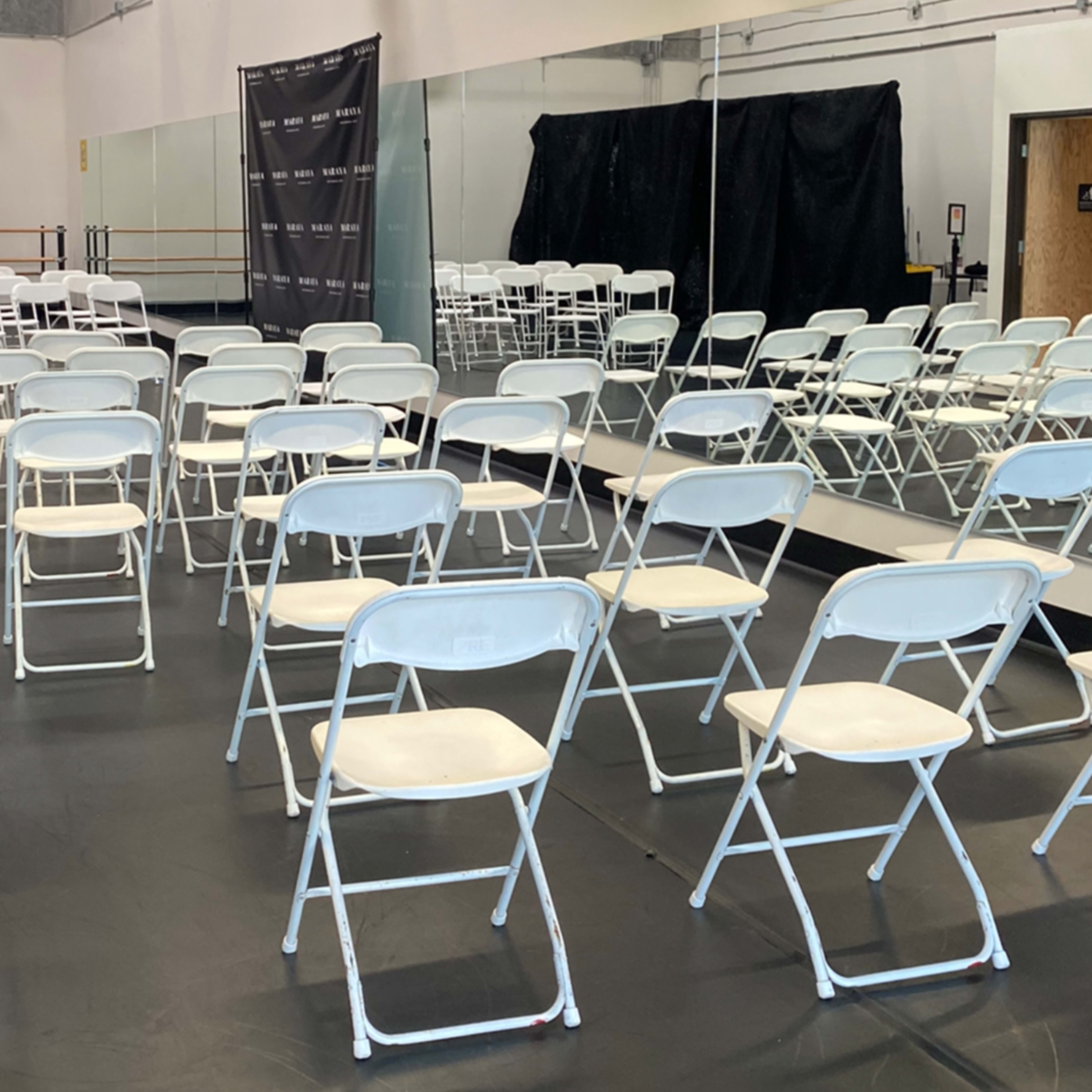 A room set up with rows of white folding chairs arranged for an event, along with mirrors on one wall and a black curtain in the back.