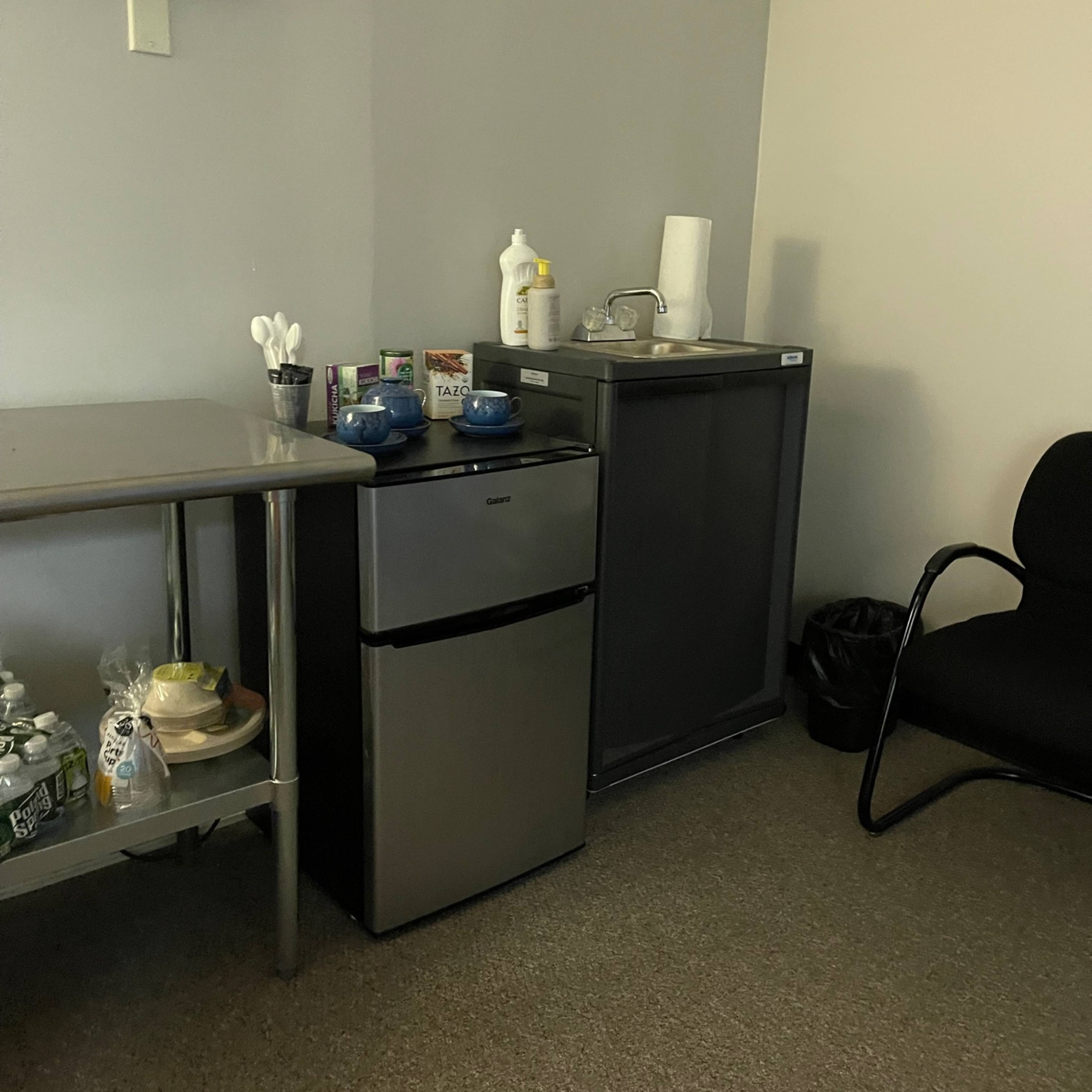 A small kitchenette area in an office with a stainless steel refrigerator, a counter with utensils and dishes, and a sink.