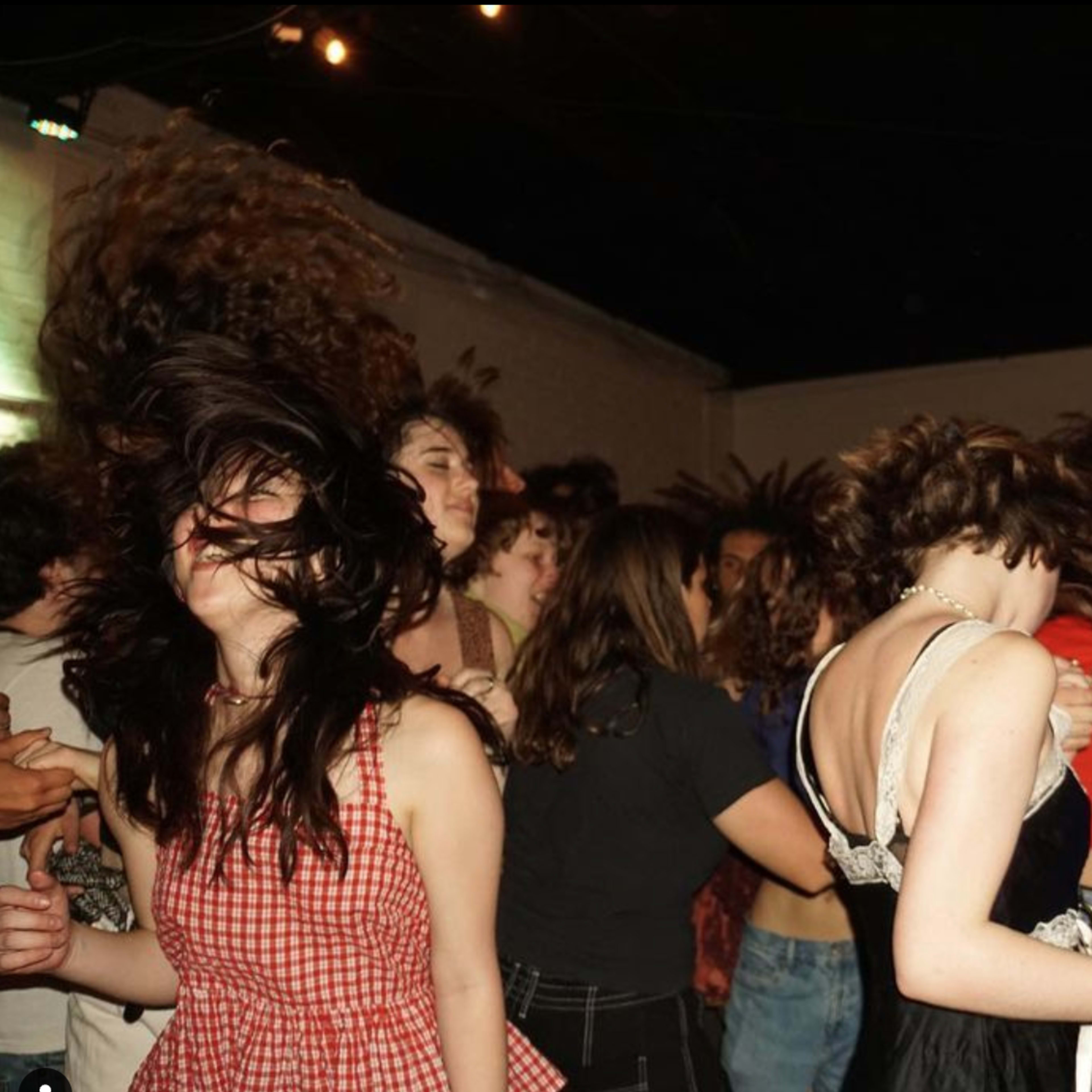 A group of young people dance energetically in a lively outdoor setting at night, with their hair moving dramatically.