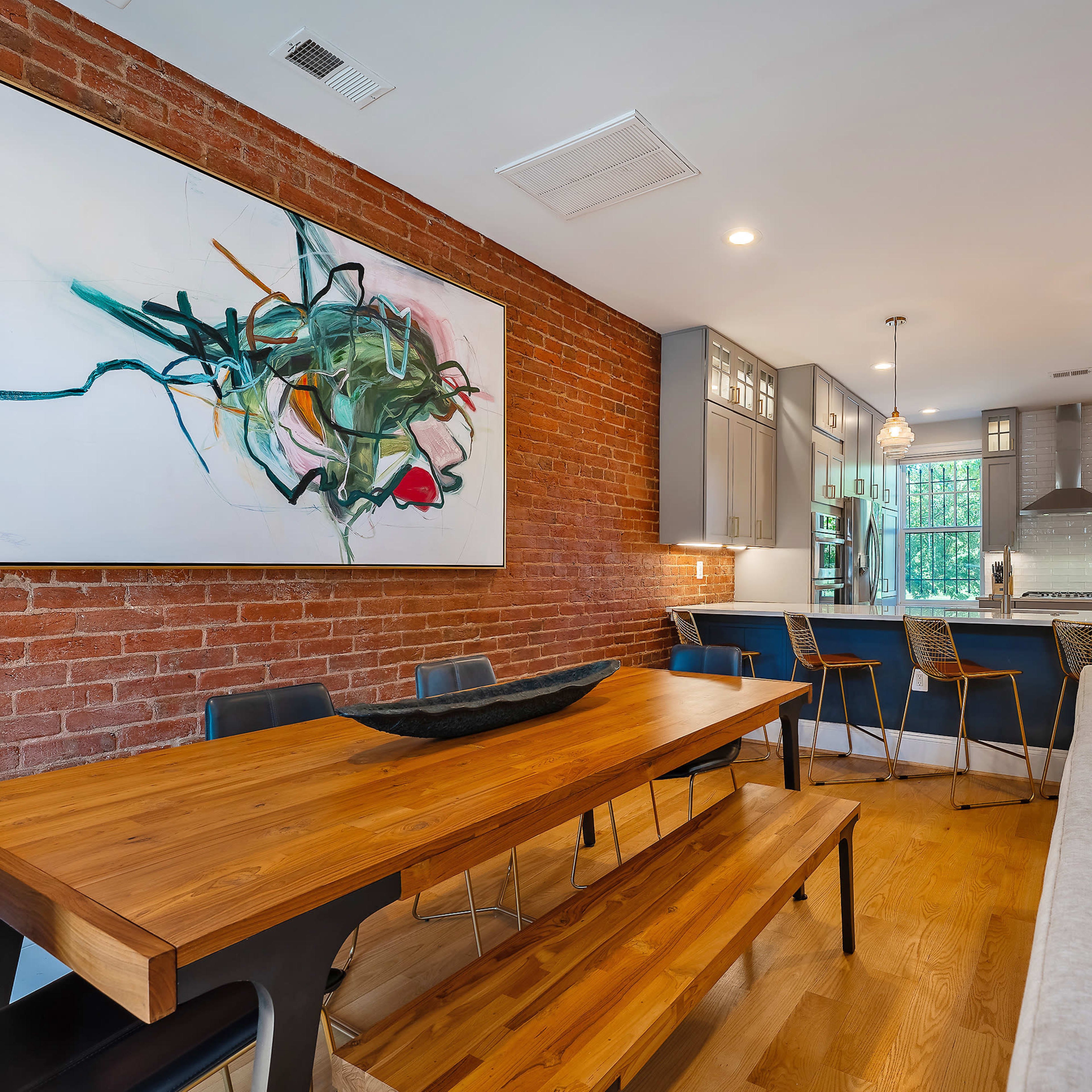 A dining area features a wooden table and bench, adjacent to a modern kitchen with gray cabinets and a large artwork on a brick wall.