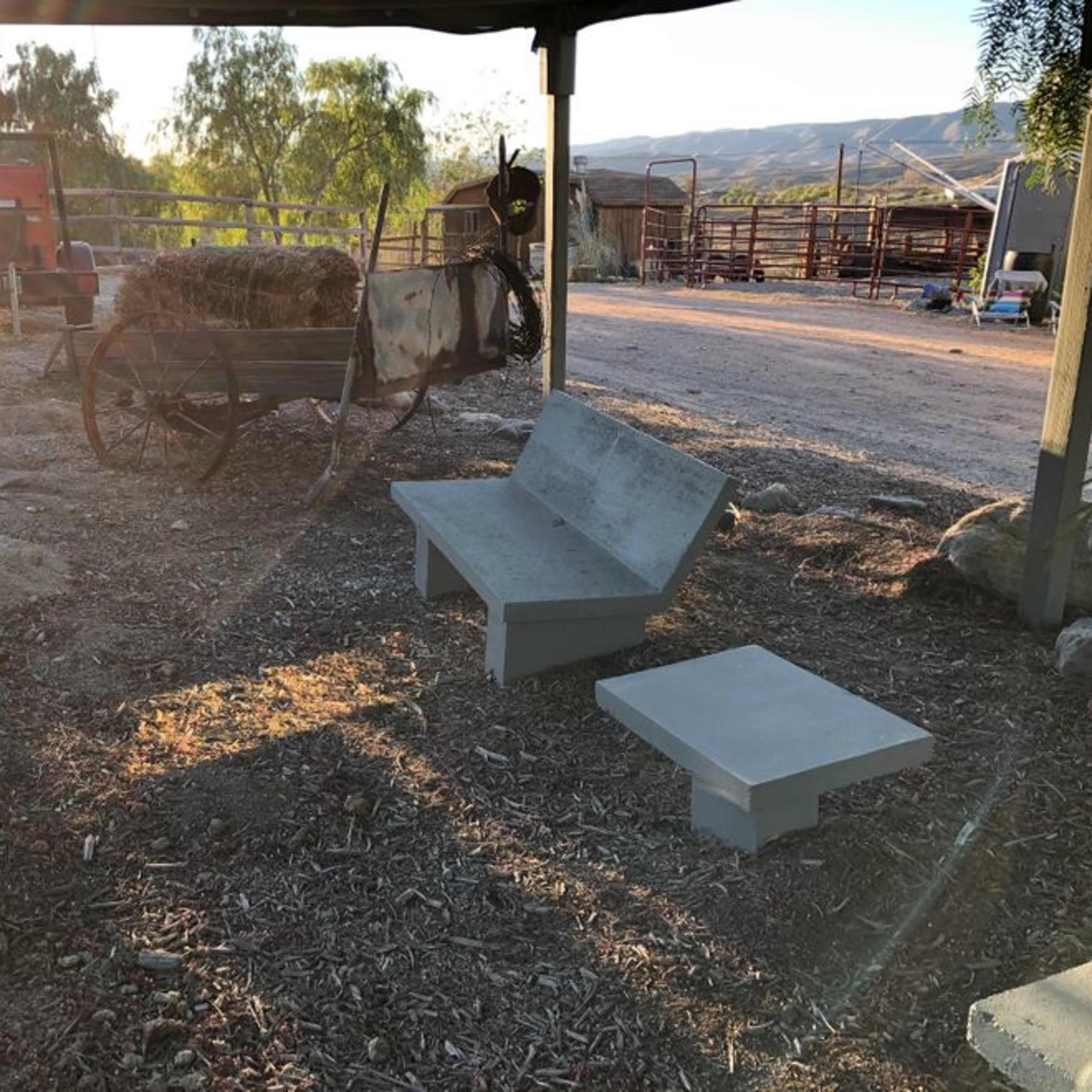 A shaded area with concrete benches, a wooden cart, and a rustic farm setting in the background.