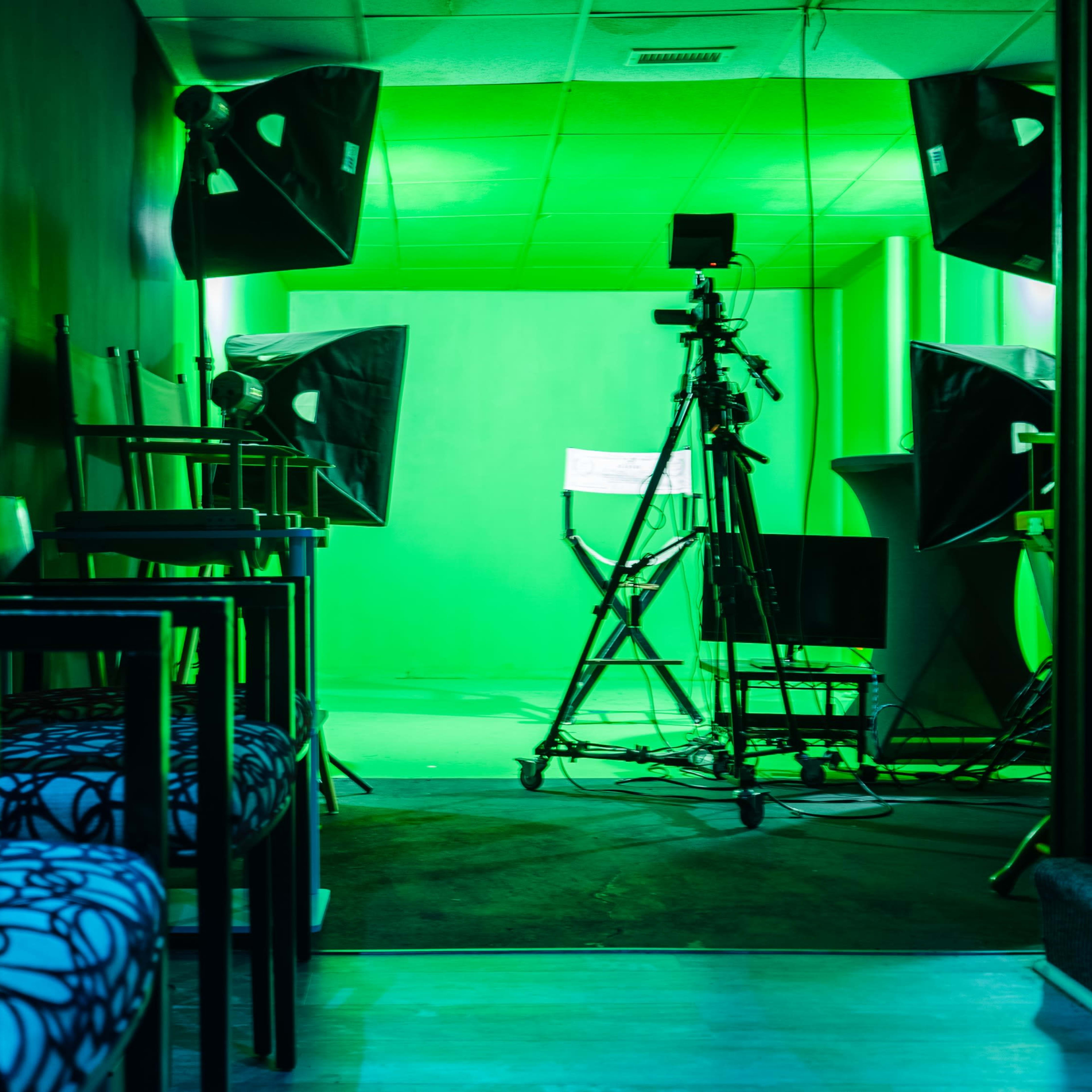 A green-lit studio space with a camera setup, lighting equipment, and chairs along the side.