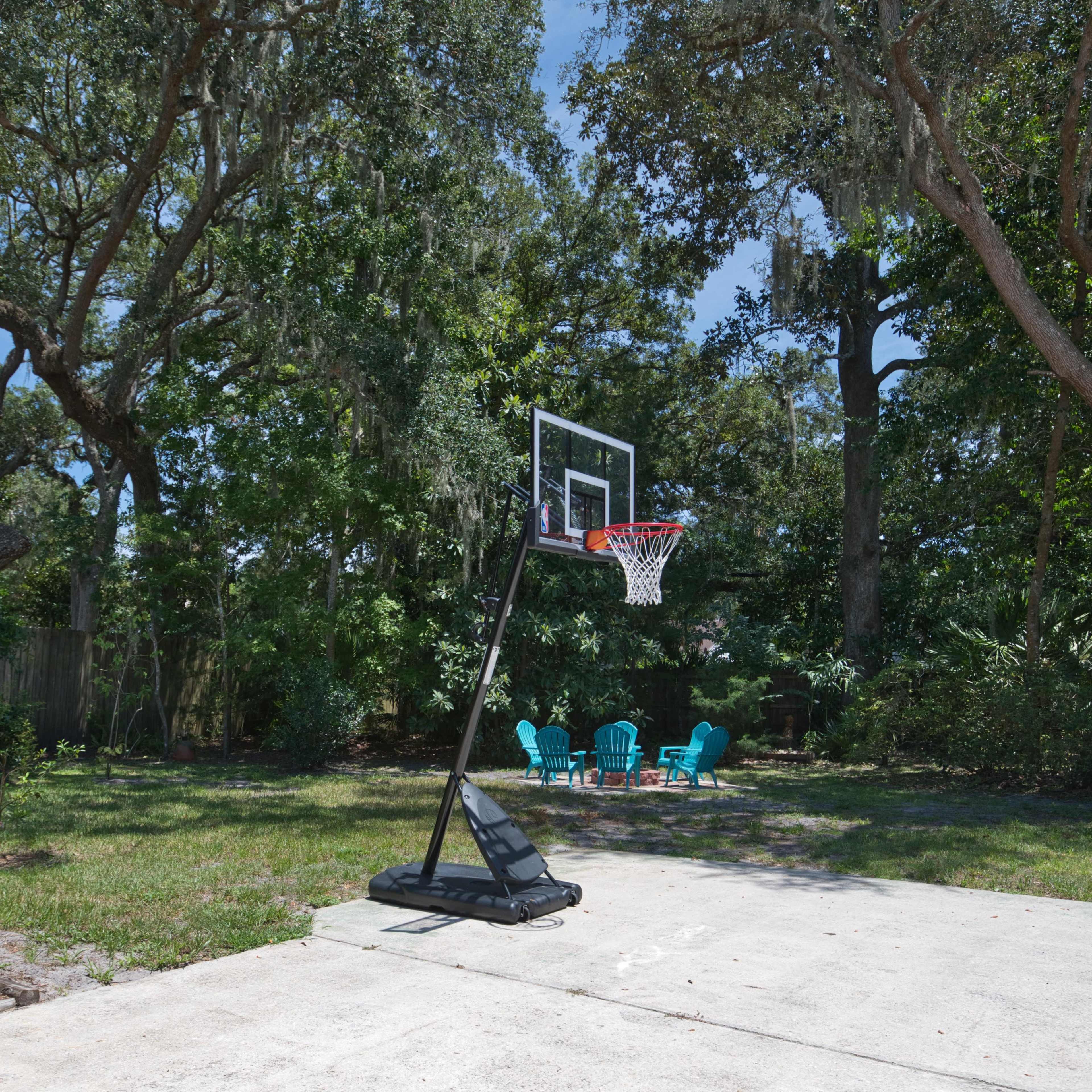 A basketball hoop stands on a paved area surrounded by grassy space and a cluster of blue chairs under large trees in a backyard.