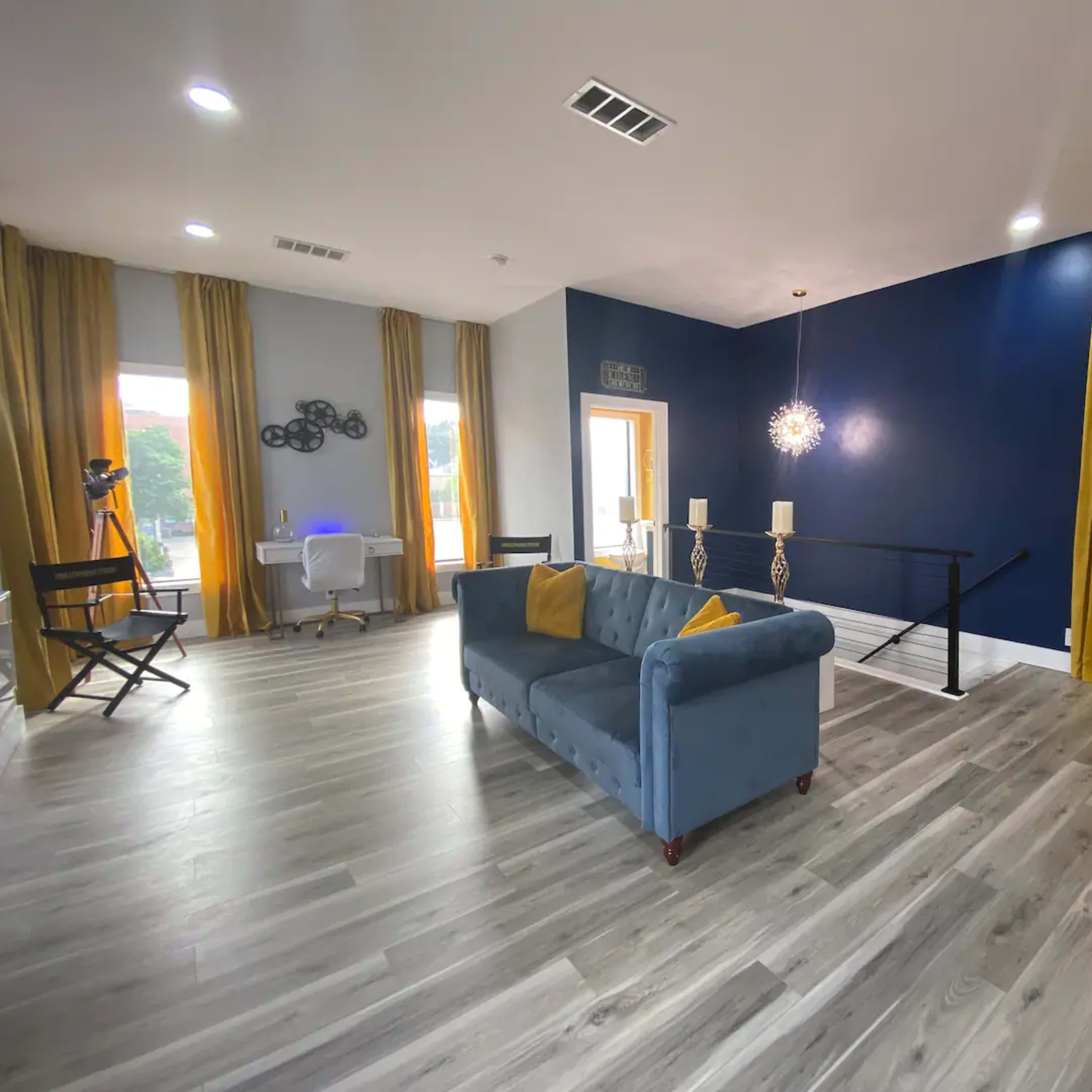 A modern living room with a gray couch, large windows with yellow curtains, and a mix of natural and artificial lighting.