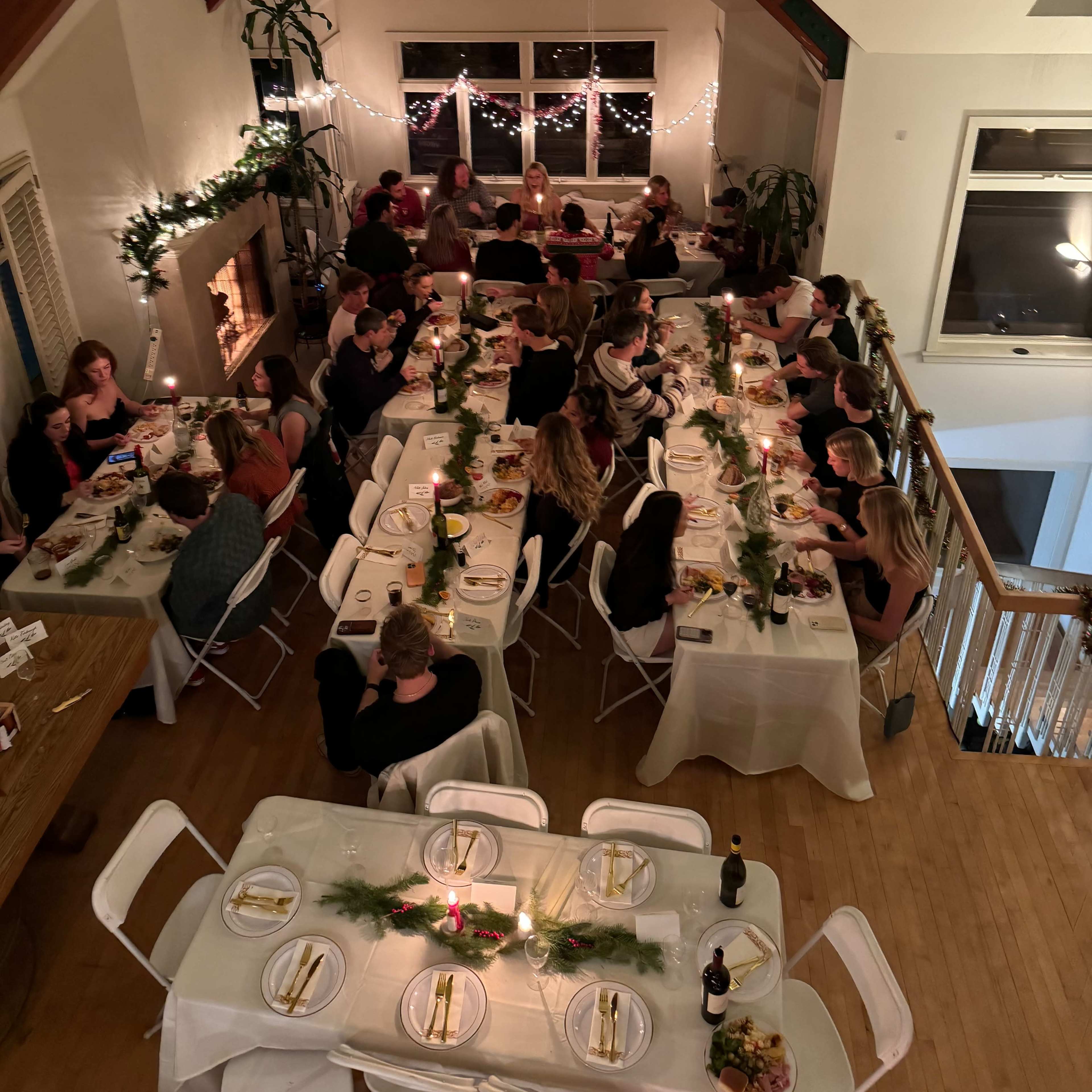 A large group of people is gathered around several long tables set for a dinner in a warmly lit room with wooden beams and decorative greenery.