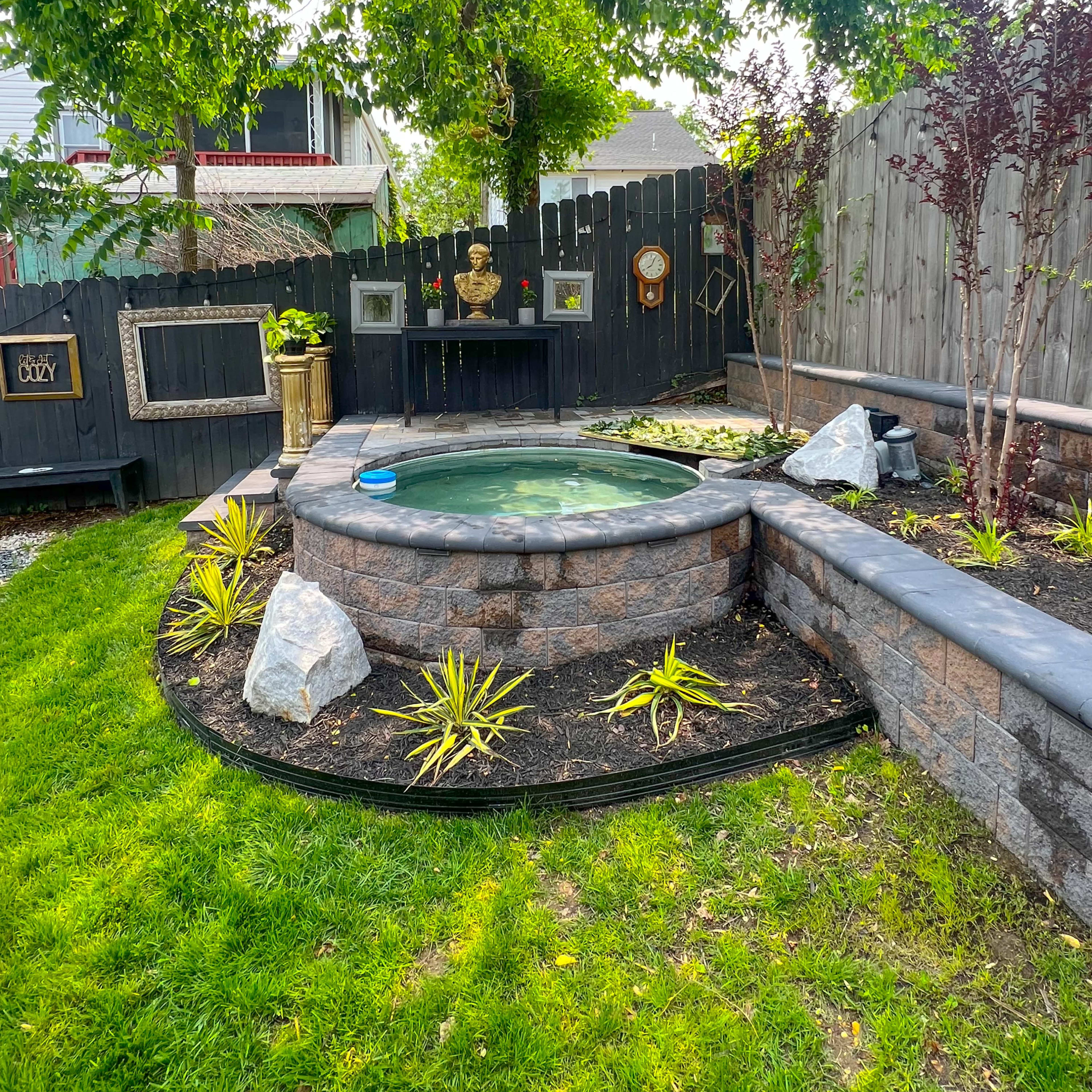 A landscaped backyard featuring a circular hot tub surrounded by stone walls, greenery, and decorative plants, with a wooden fence and various garden elements in the background.