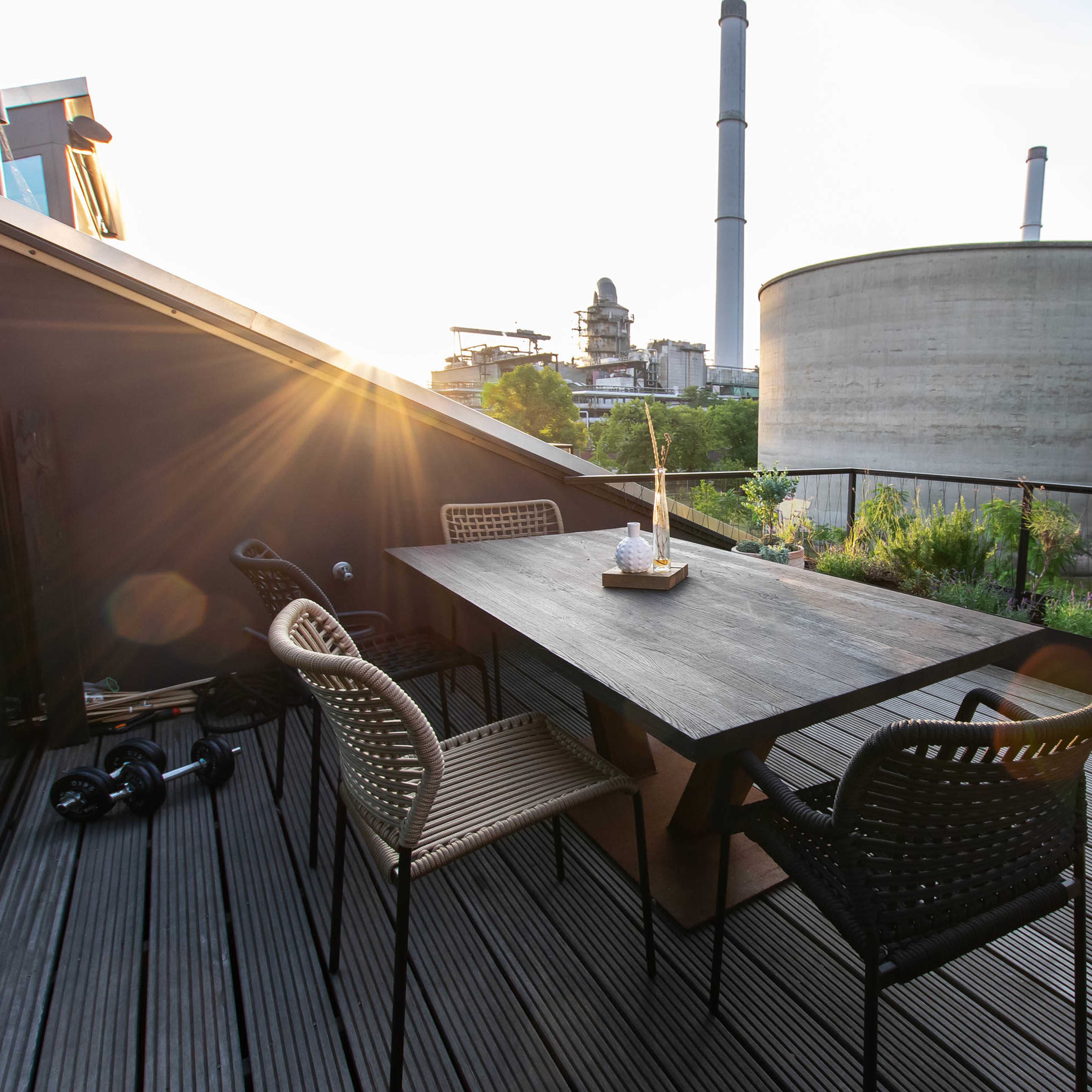 A rooftop terrace features a wooden dining table surrounded by chairs, overlooking industrial buildings and greenery at sunset.