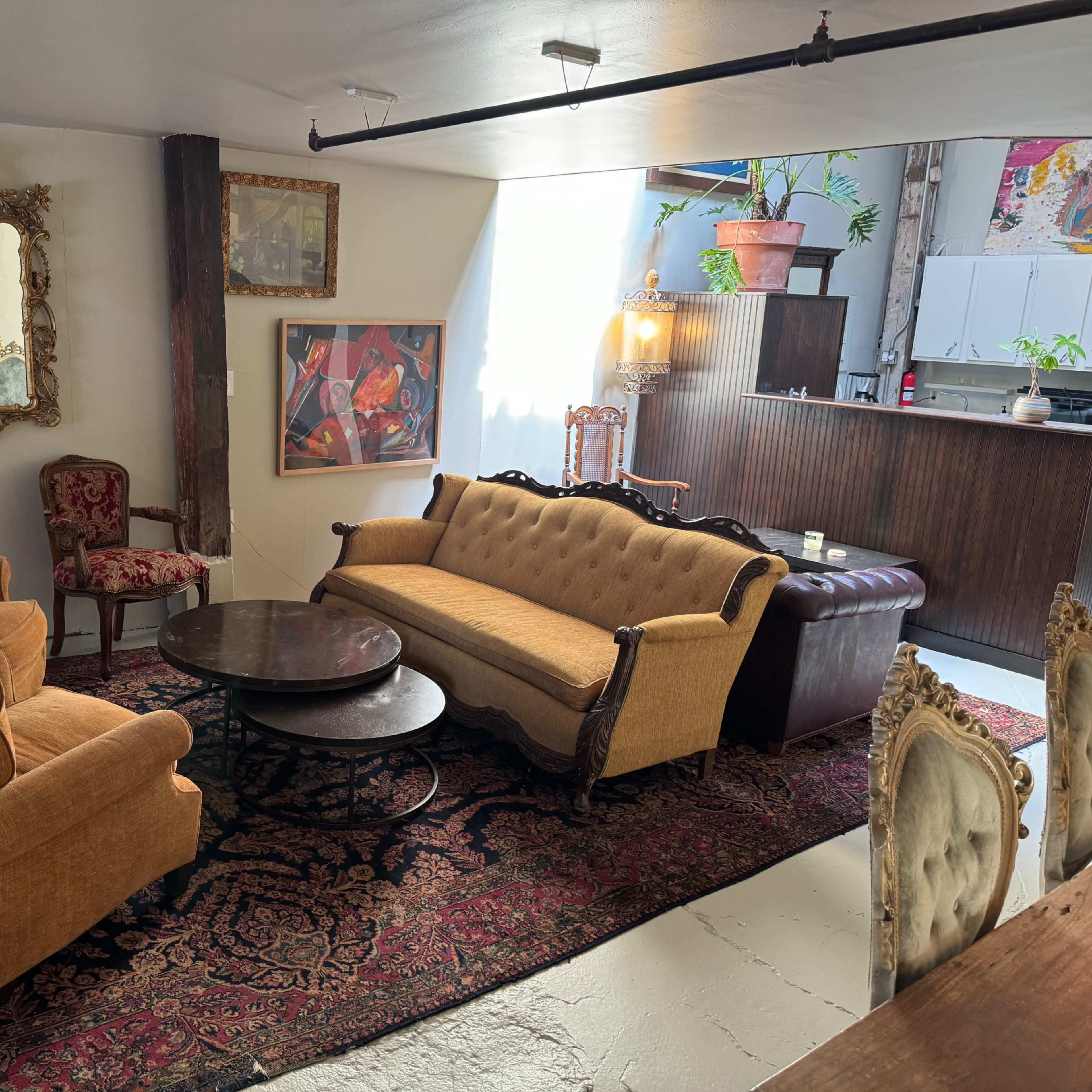 A cozy living room featuring a vintage-style sofa, two armchairs, a round coffee table, and an area rug, with a kitchenette visible in the background.