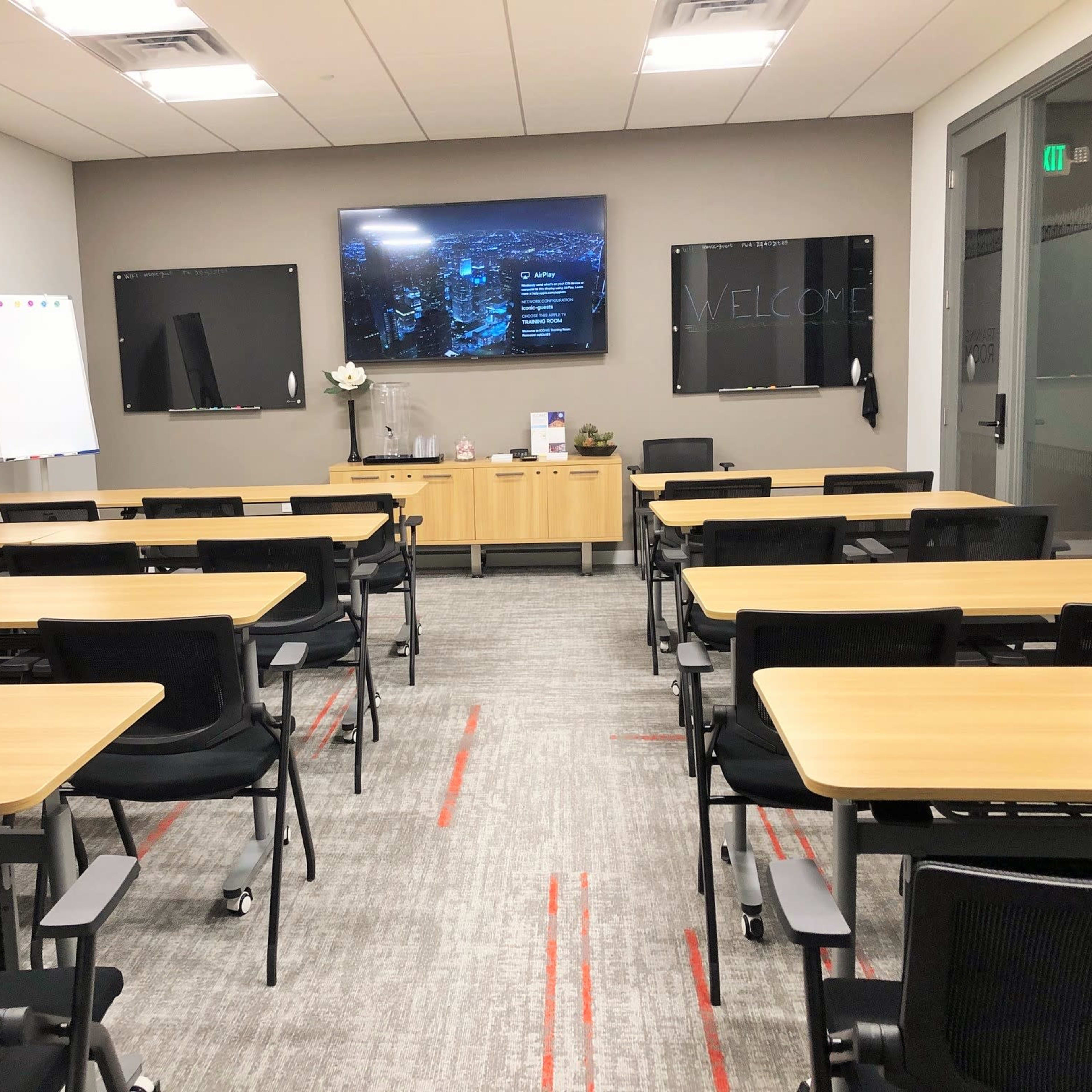 A modern meeting room is set up with several rectangular tables and chairs arranged facing a wall-mounted screen displaying a welcome message.