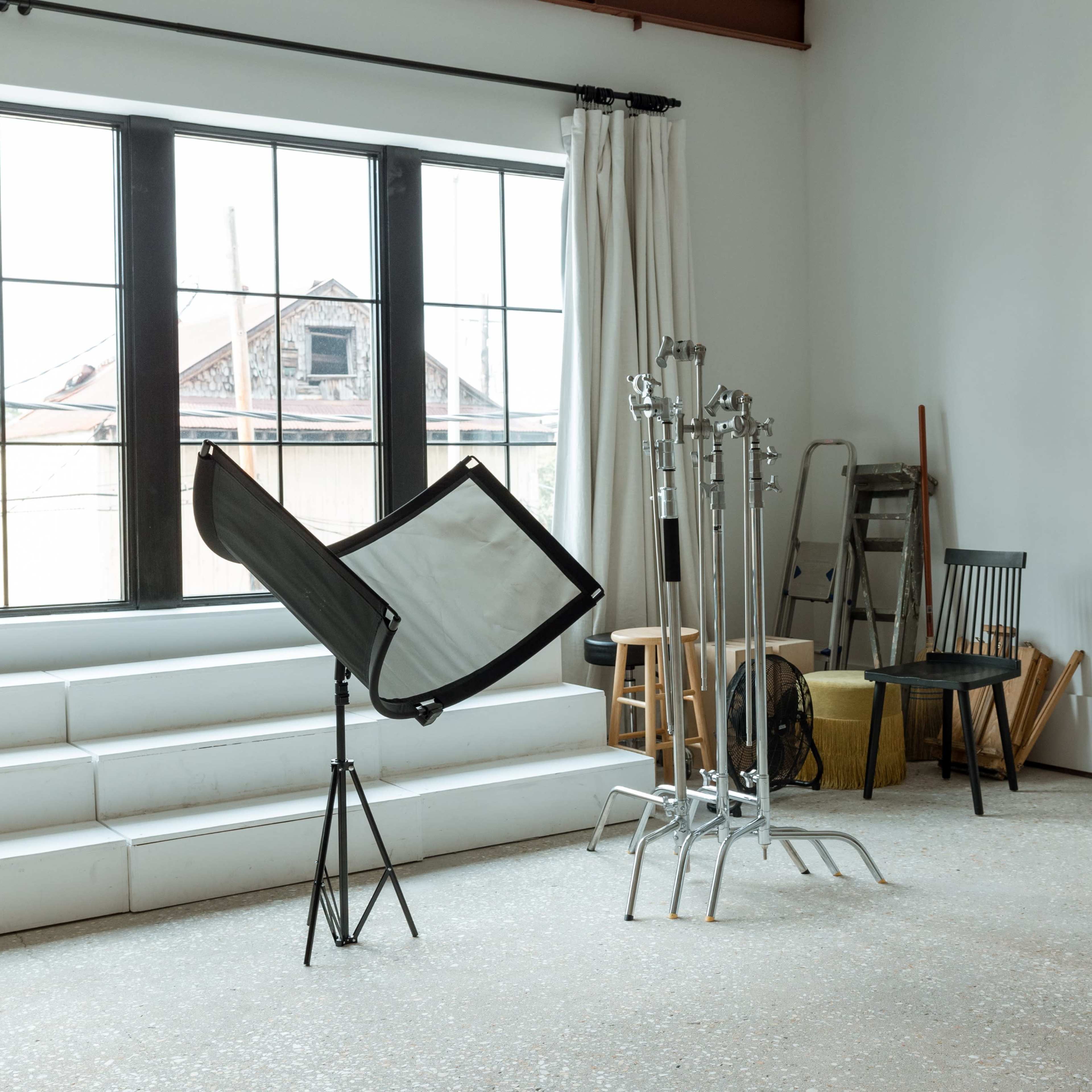 A photography studio with a large window, a light reflector on a stand, and several light stands along with a chair and a ladder in the corner.