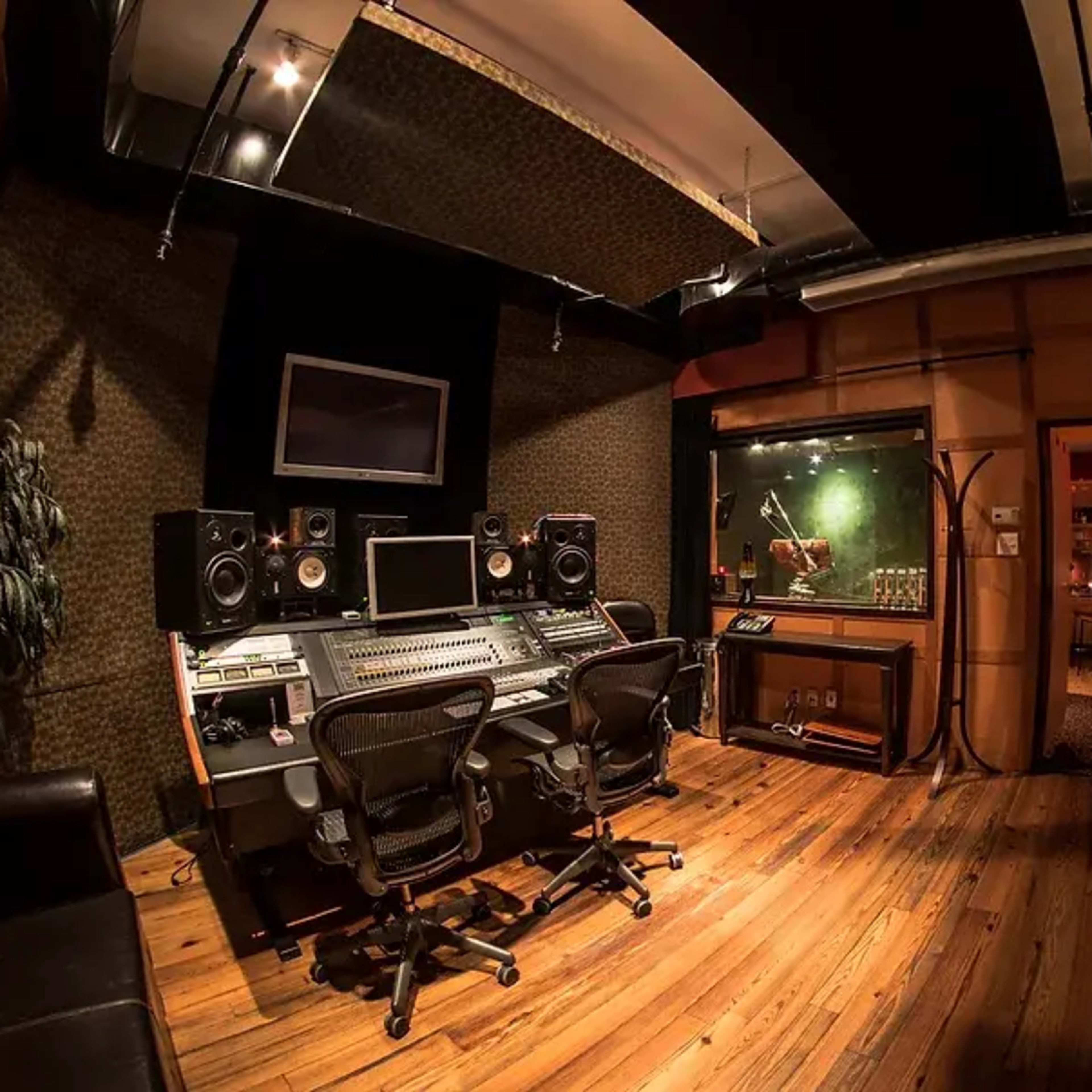 A soundproof recording studio featuring a large mixing console, two studio monitors, and a glass vocal booth alongside leather chairs and wooden flooring.