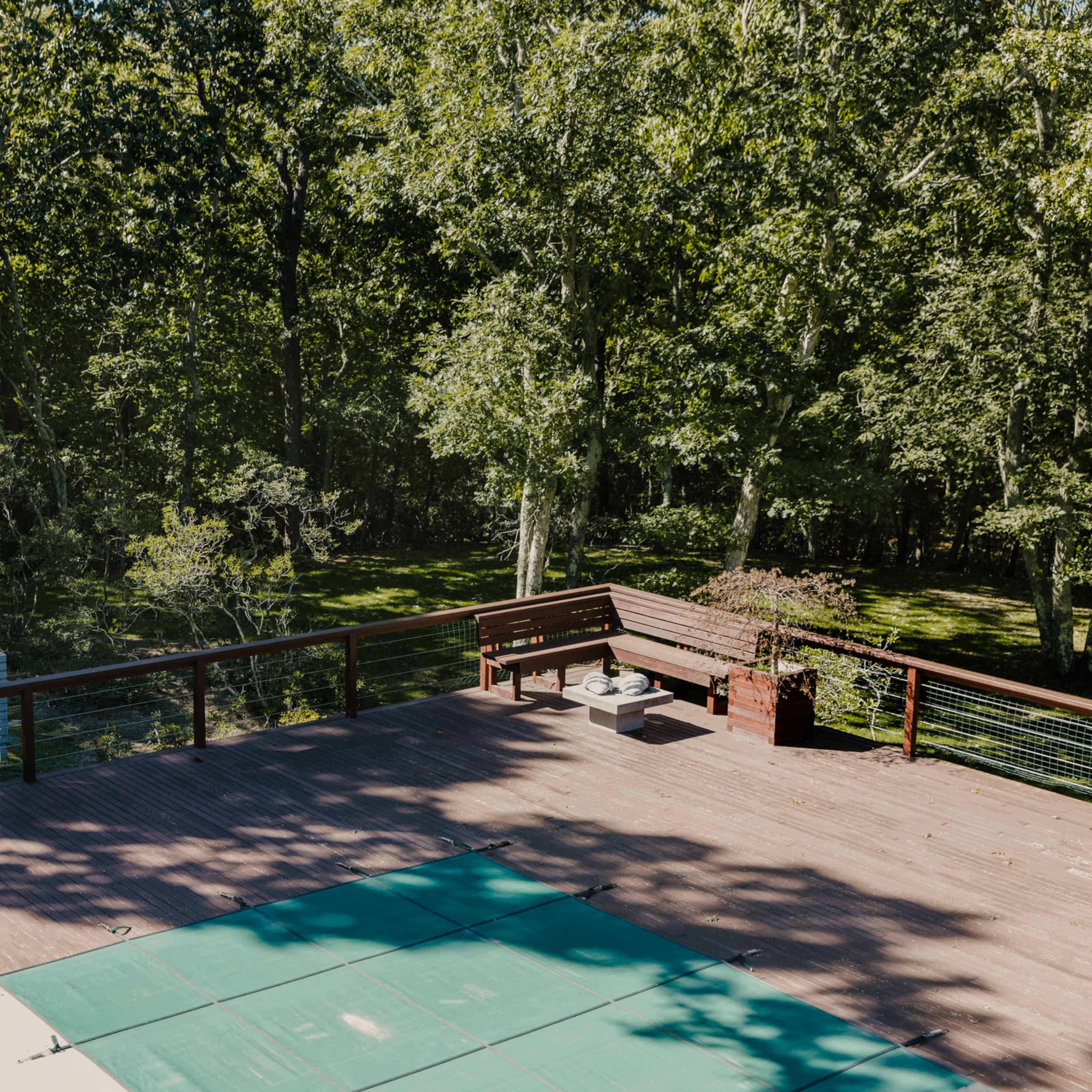 A wooden deck surrounded by trees, featuring a green pool cover and a bench at one end.