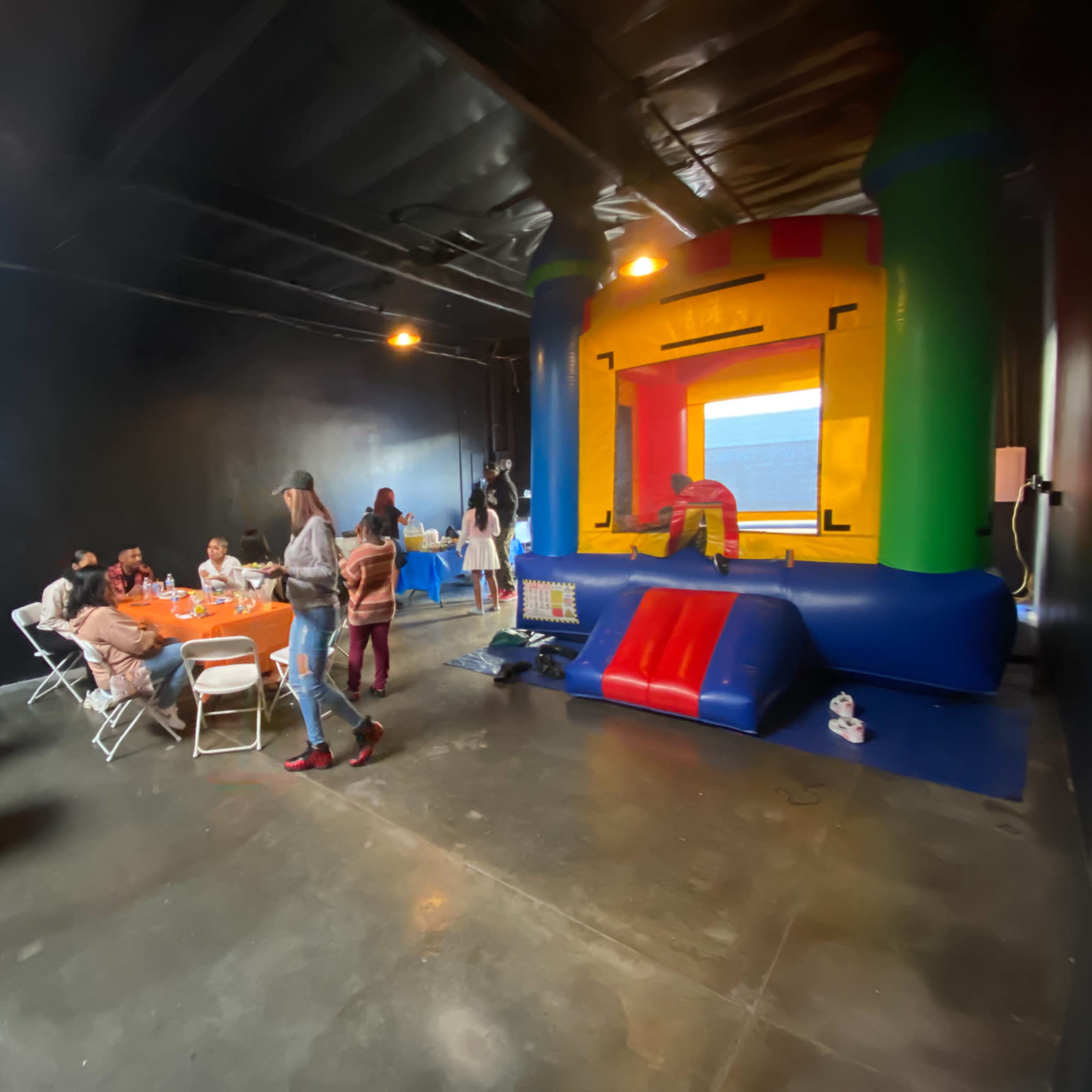 A group of people sits at tables while a colorful inflatable bounce house stands in the corner of a dimly lit room.