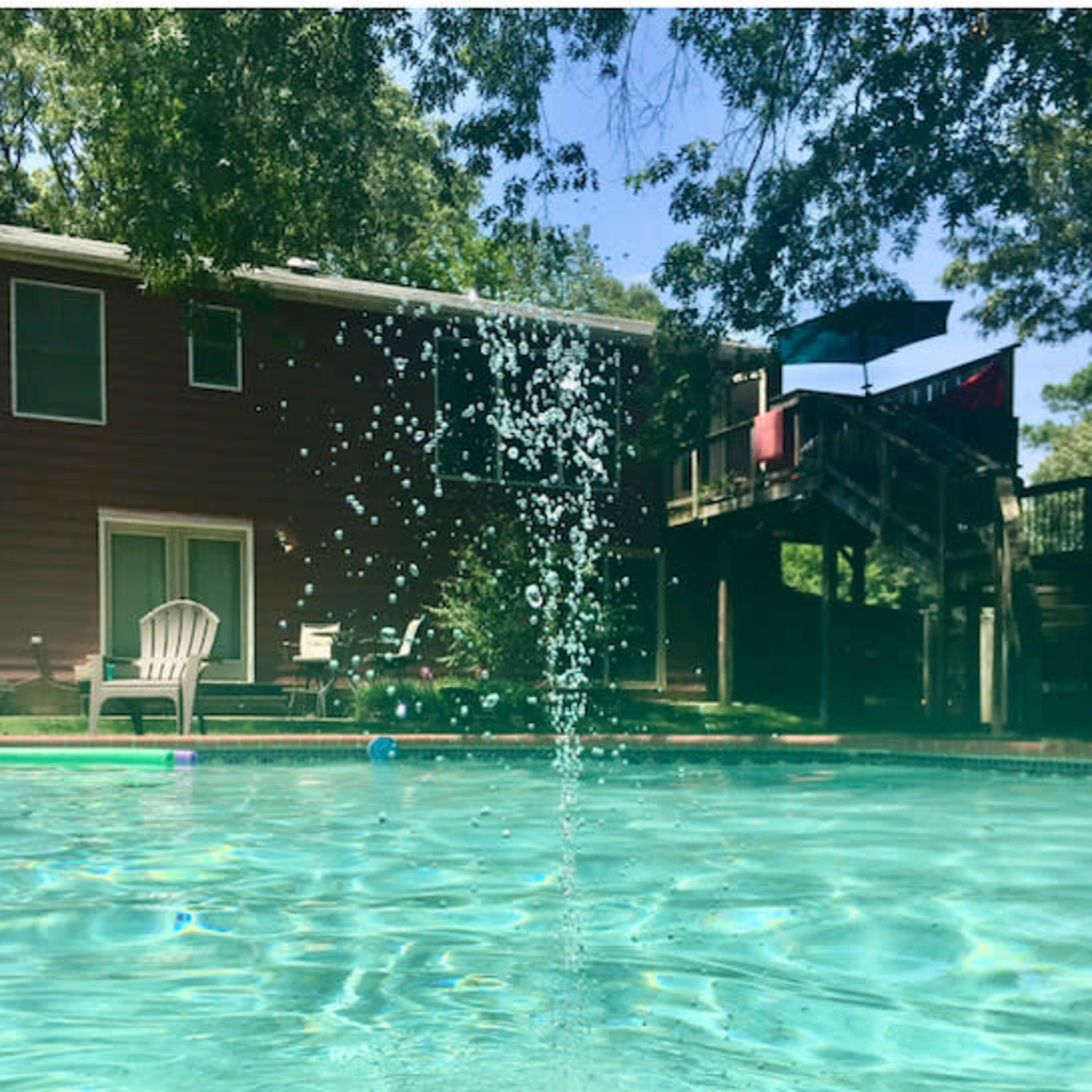 A fountain sprays water into a swimming pool, with a backyard and house visible in the background.