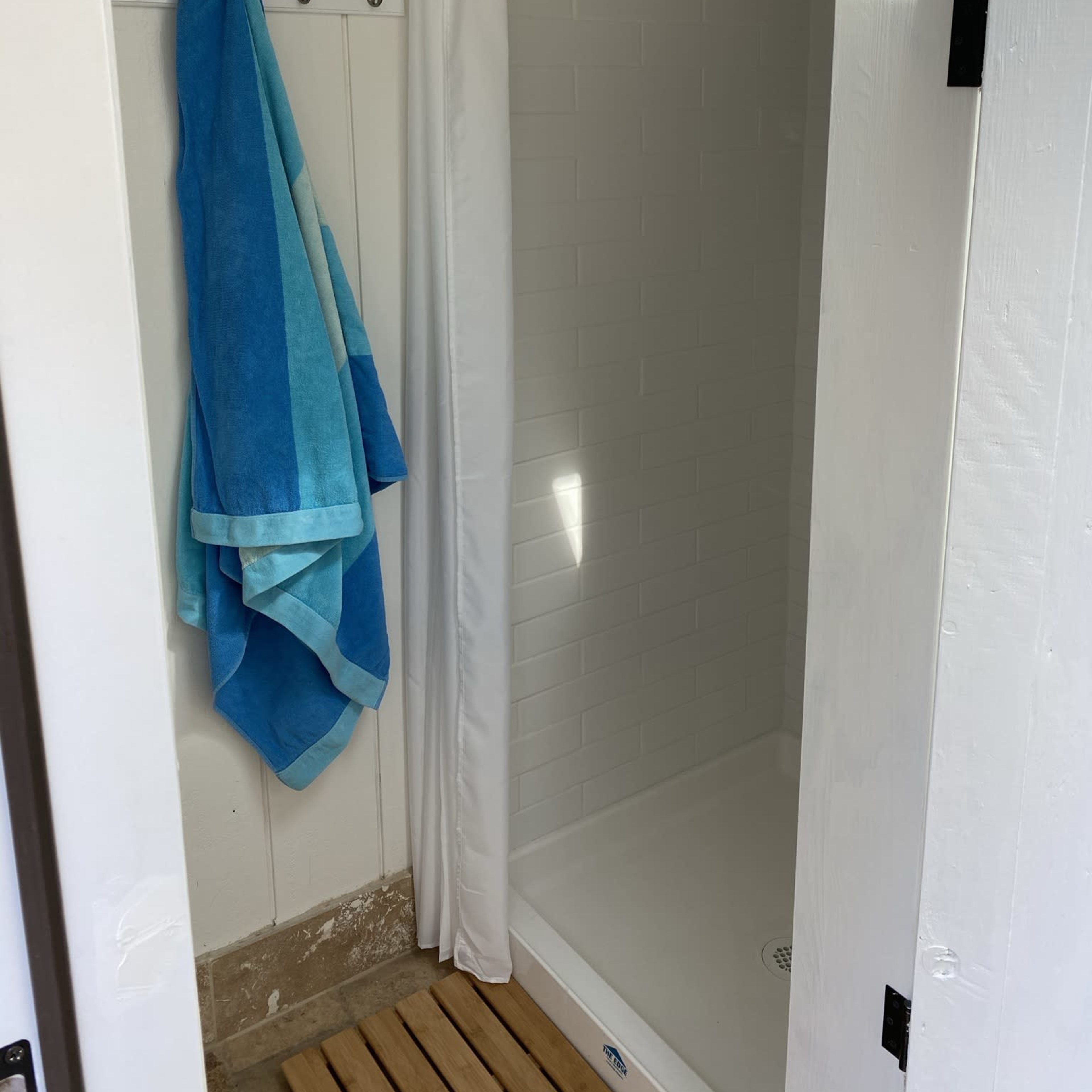 A shower area with a white curtain, a light blue and teal towel hung on a hook, and wooden slats on the floor.