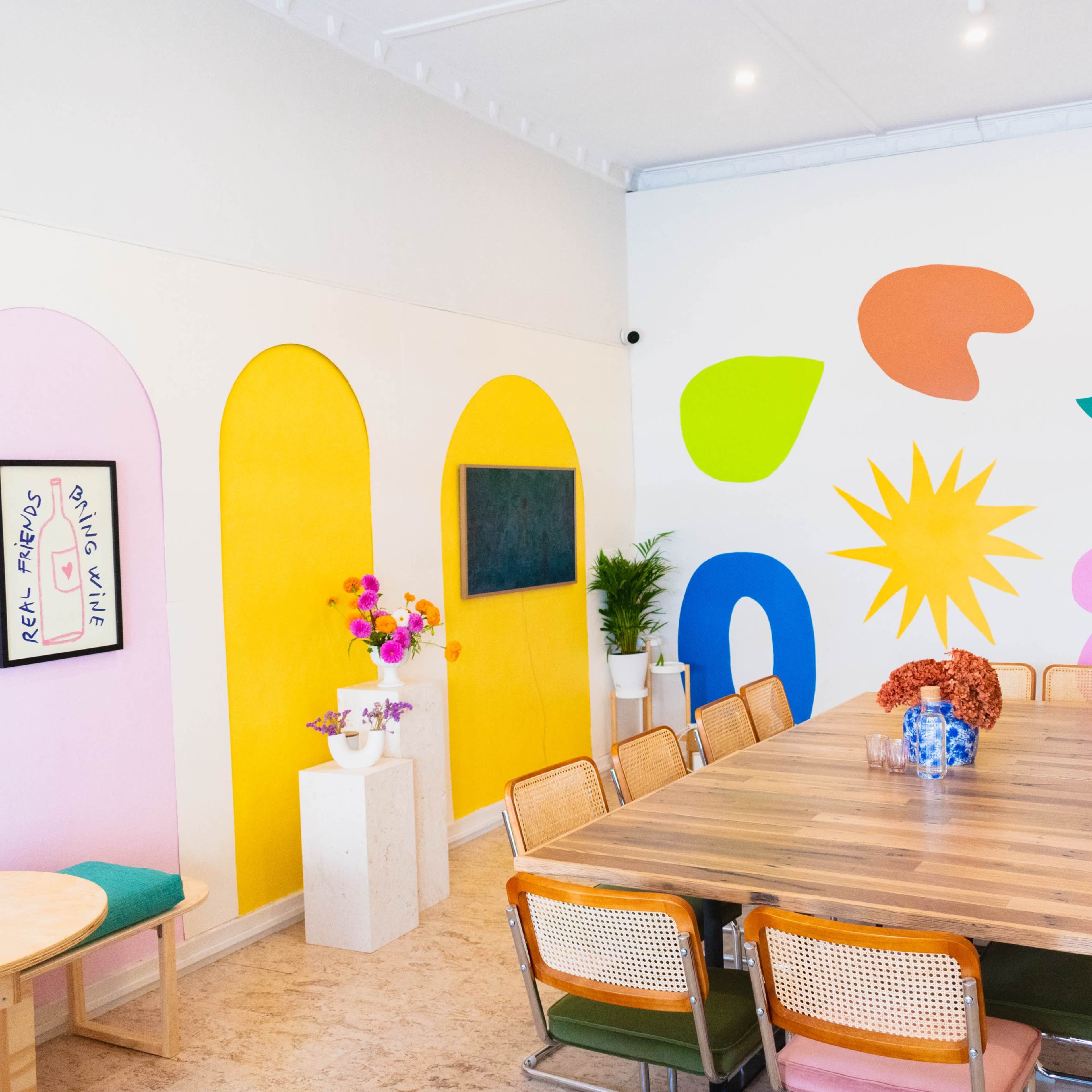 A vibrant meeting room featuring colorful wall art, a large wooden table surrounded by chairs, and decorative plants.