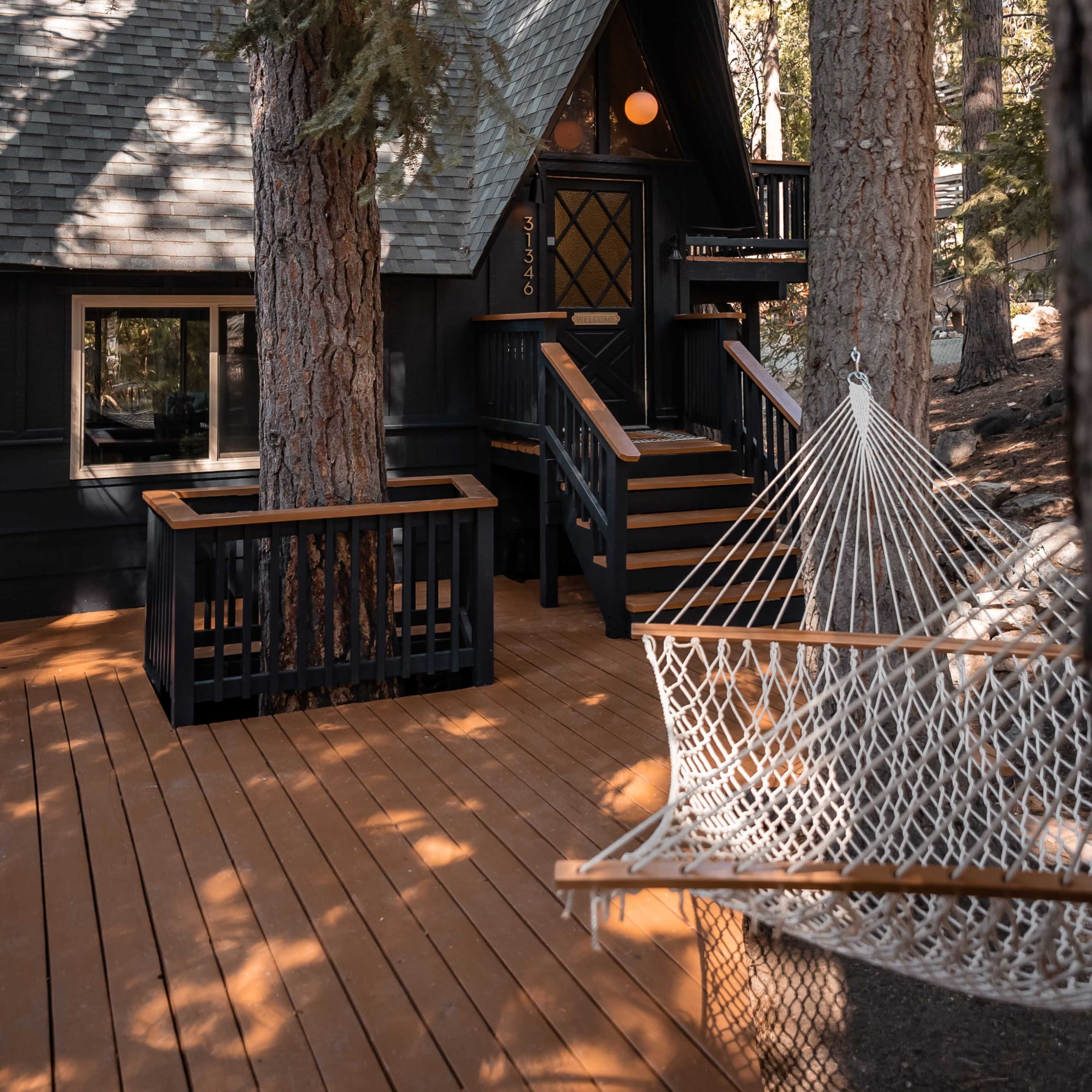 A dark wooden cabin with steep rooflines is nestled among tall trees, featuring a wooden deck and a hammock nearby.