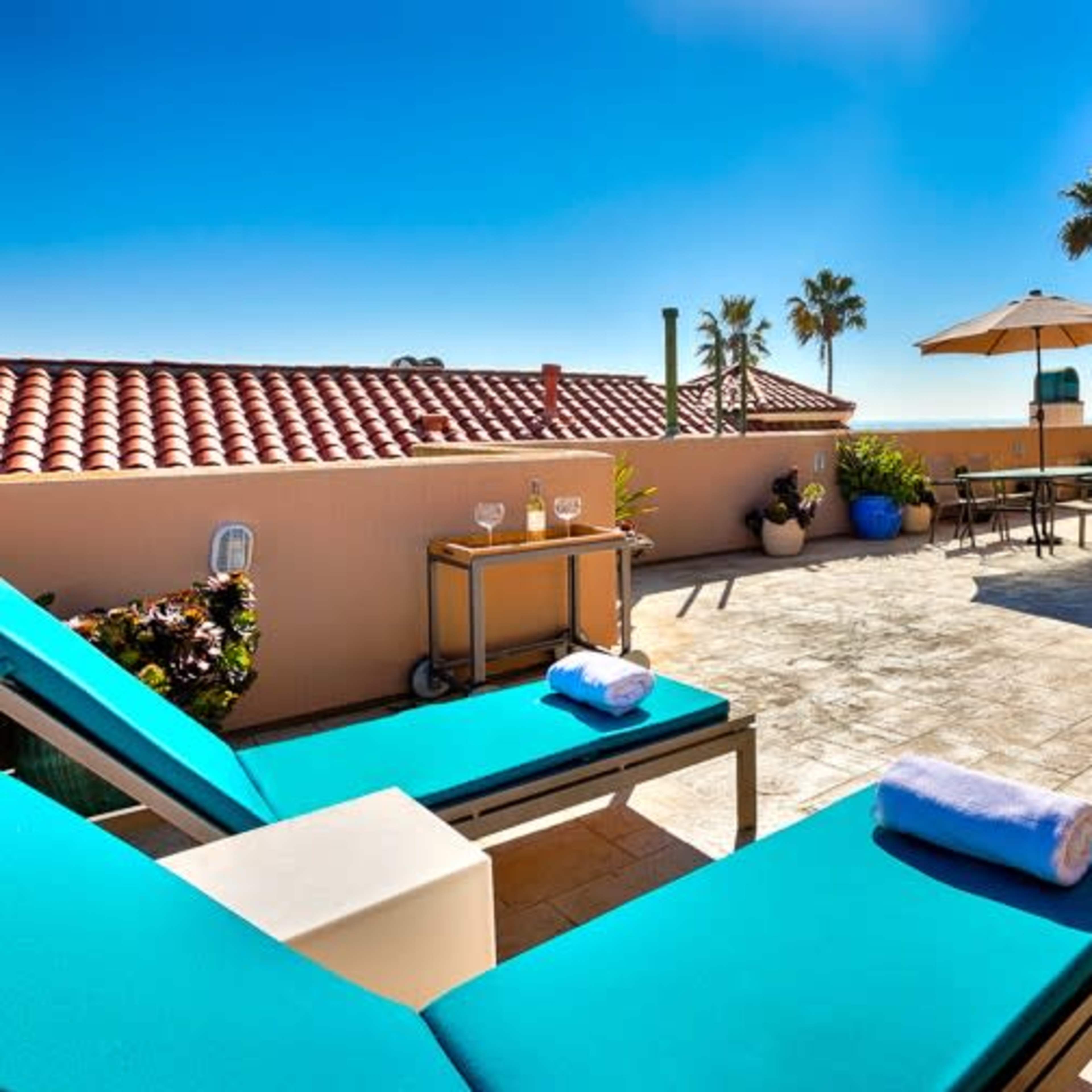 A sunlit outdoor patio with turquoise lounge chairs, an umbrella, and a view of the ocean.