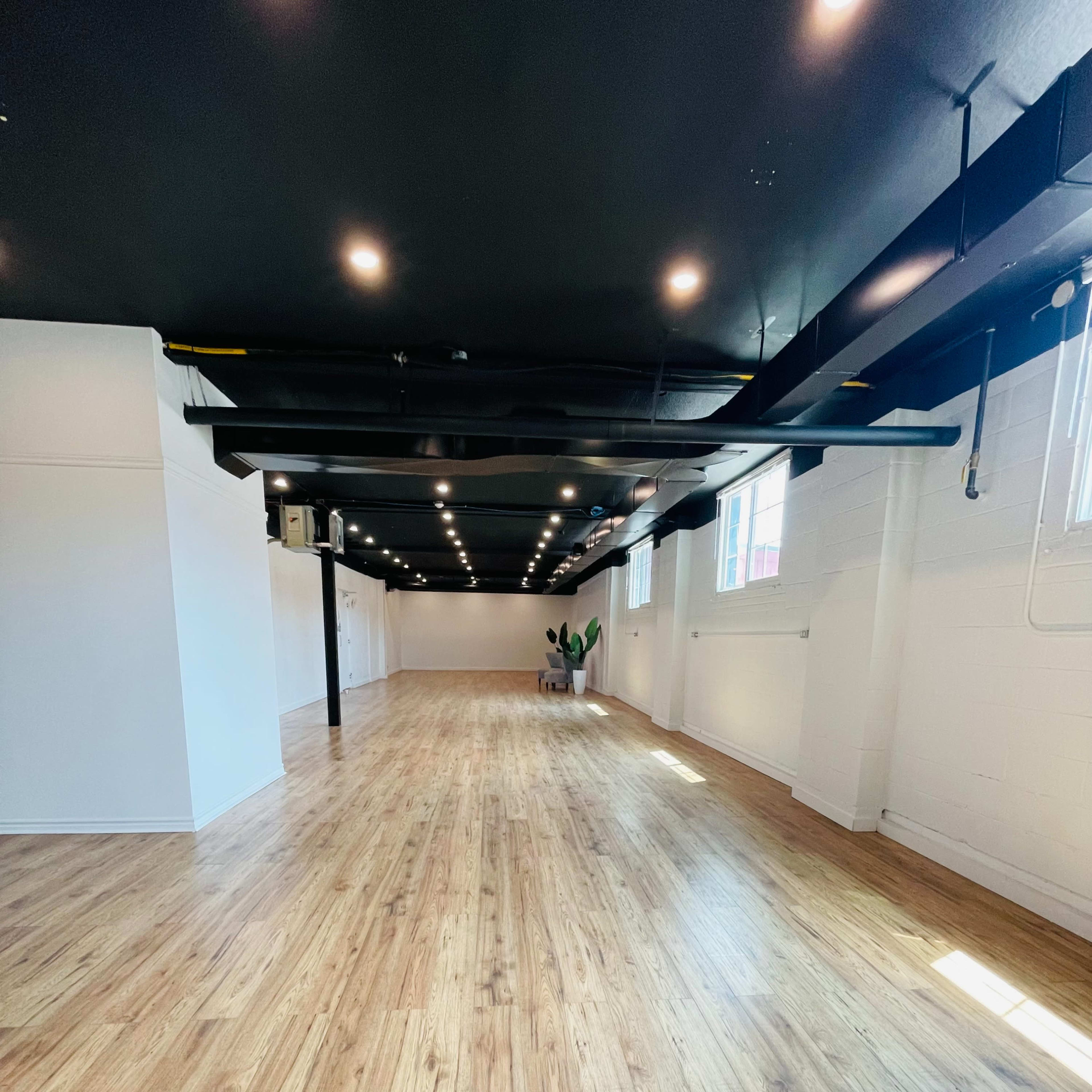 A spacious, empty room with wooden floors and black ceiling beams, featuring large windows that allow natural light to enter.