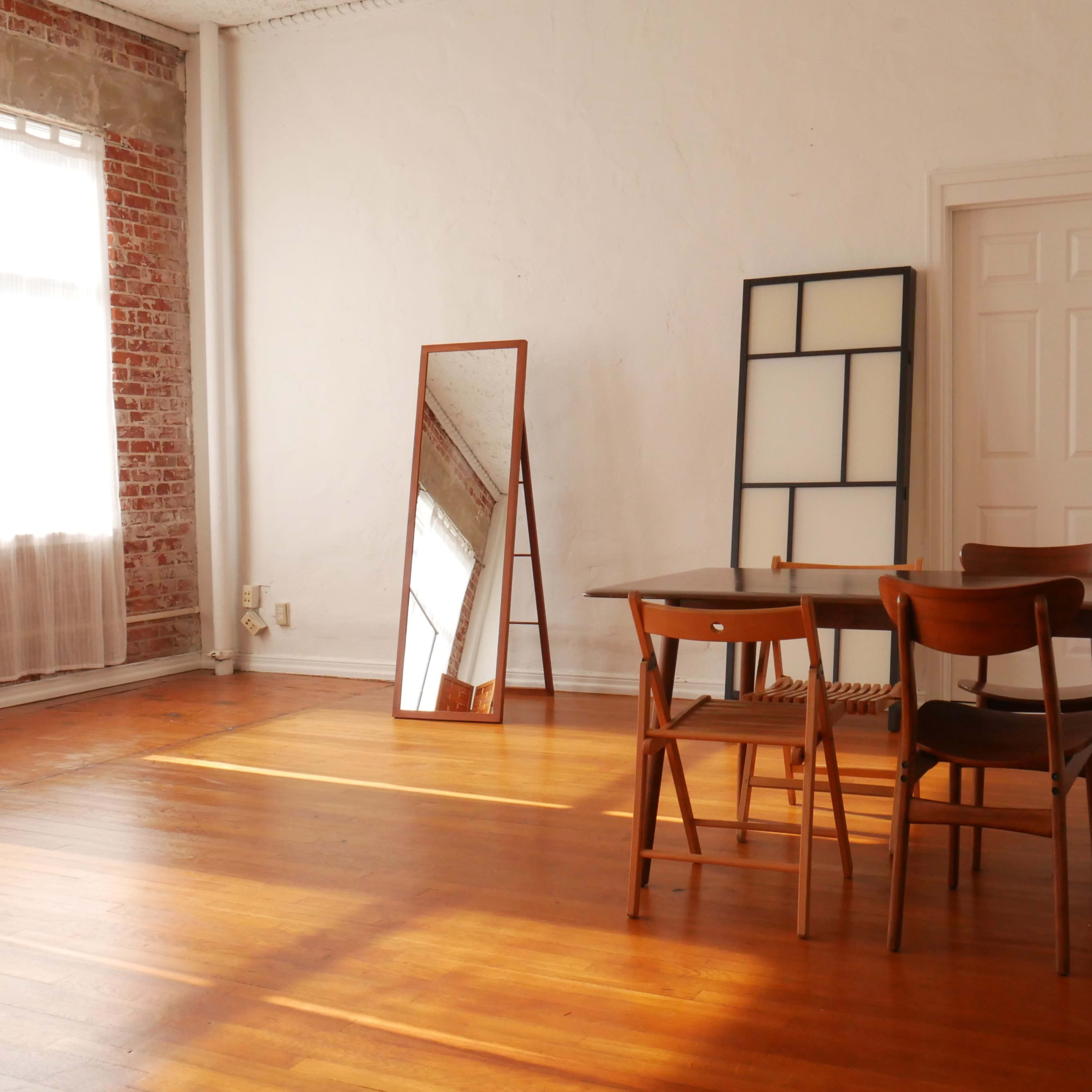 A well-lit room features a wooden table surrounded by chairs, a large mirror, and a door, with exposed brick walls and wooden flooring.