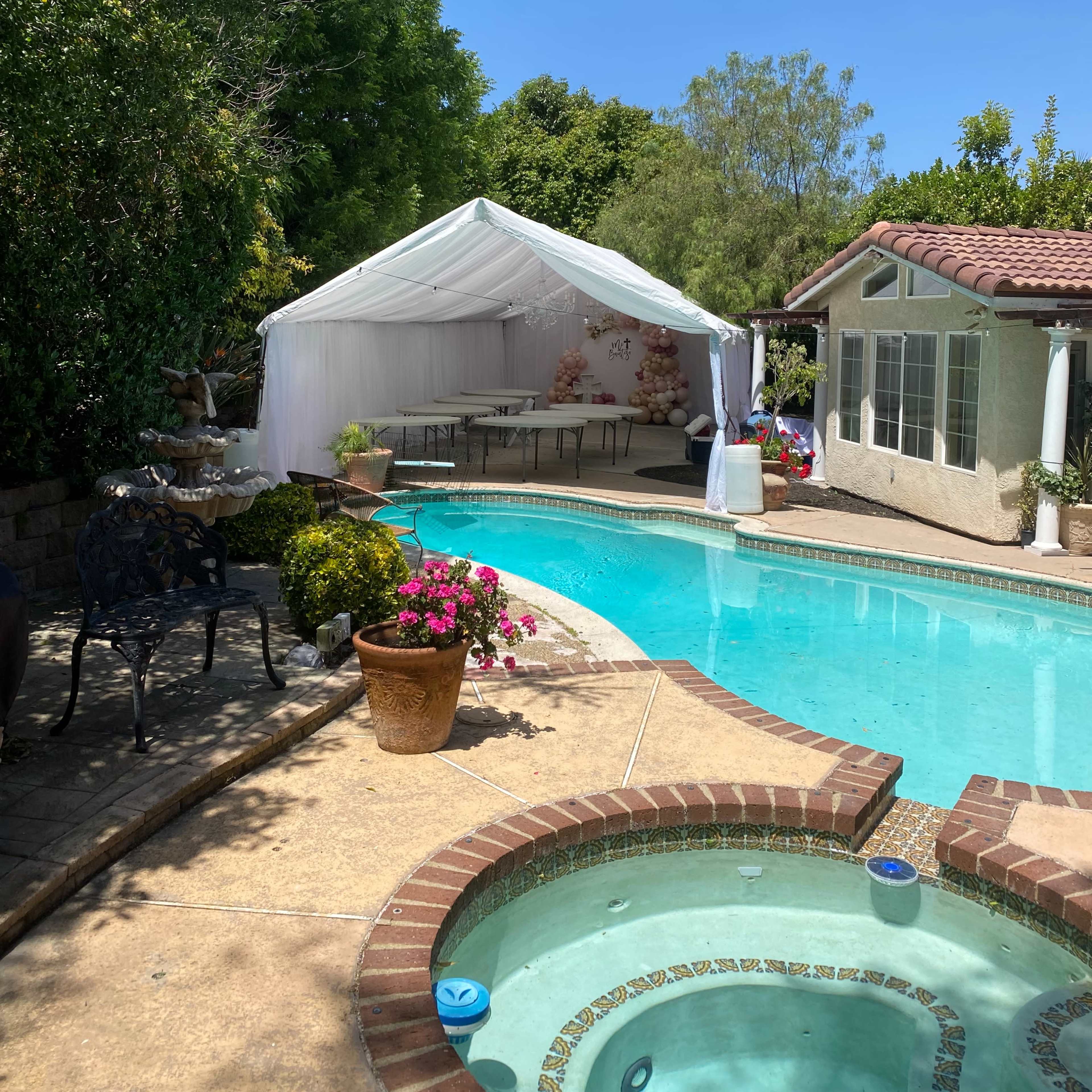 A white tent is setup beside a swimming pool in a backyard surrounded by greenery.