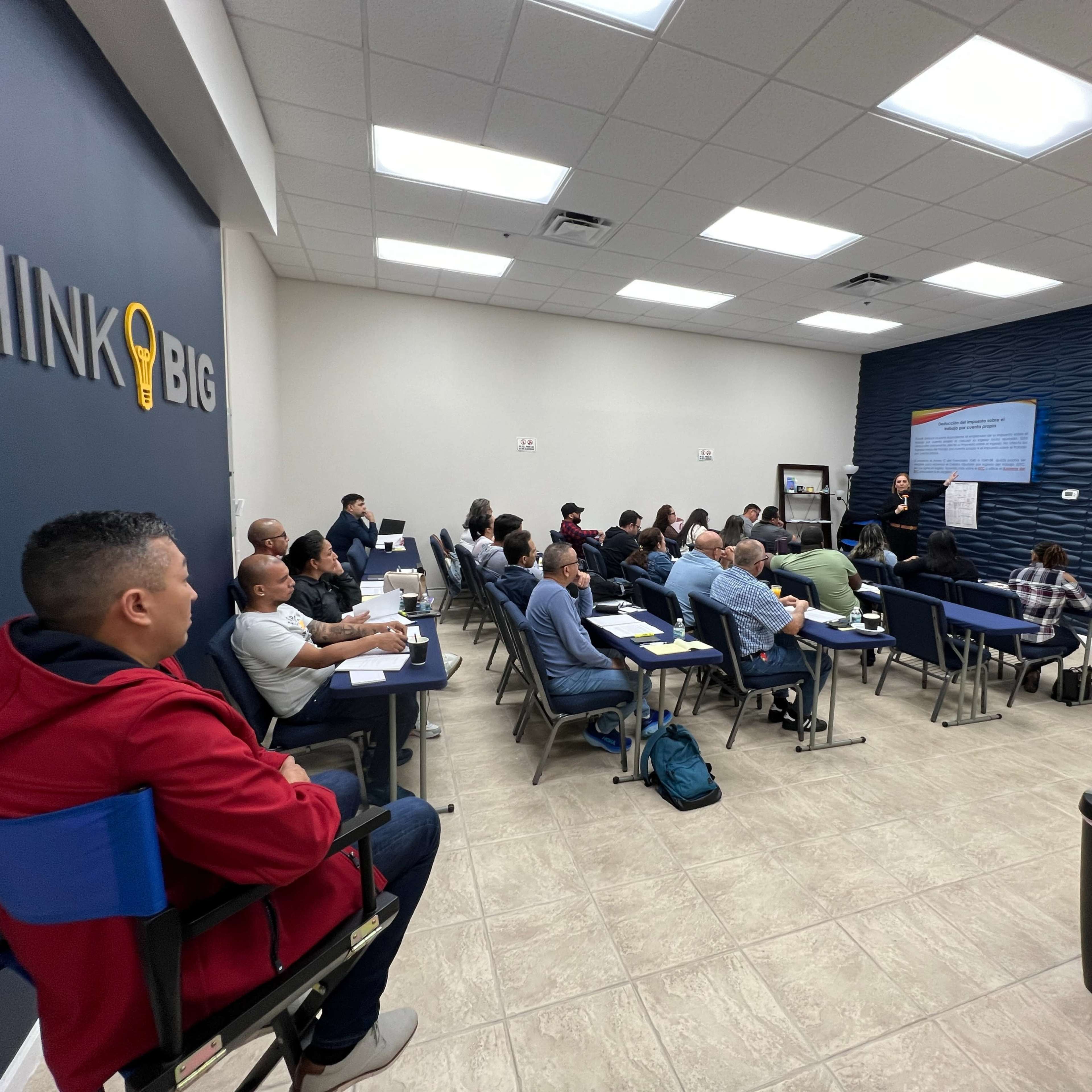 A group of individuals is seated at tables in a training room, listening to a speaker presenting in front of a large screen.