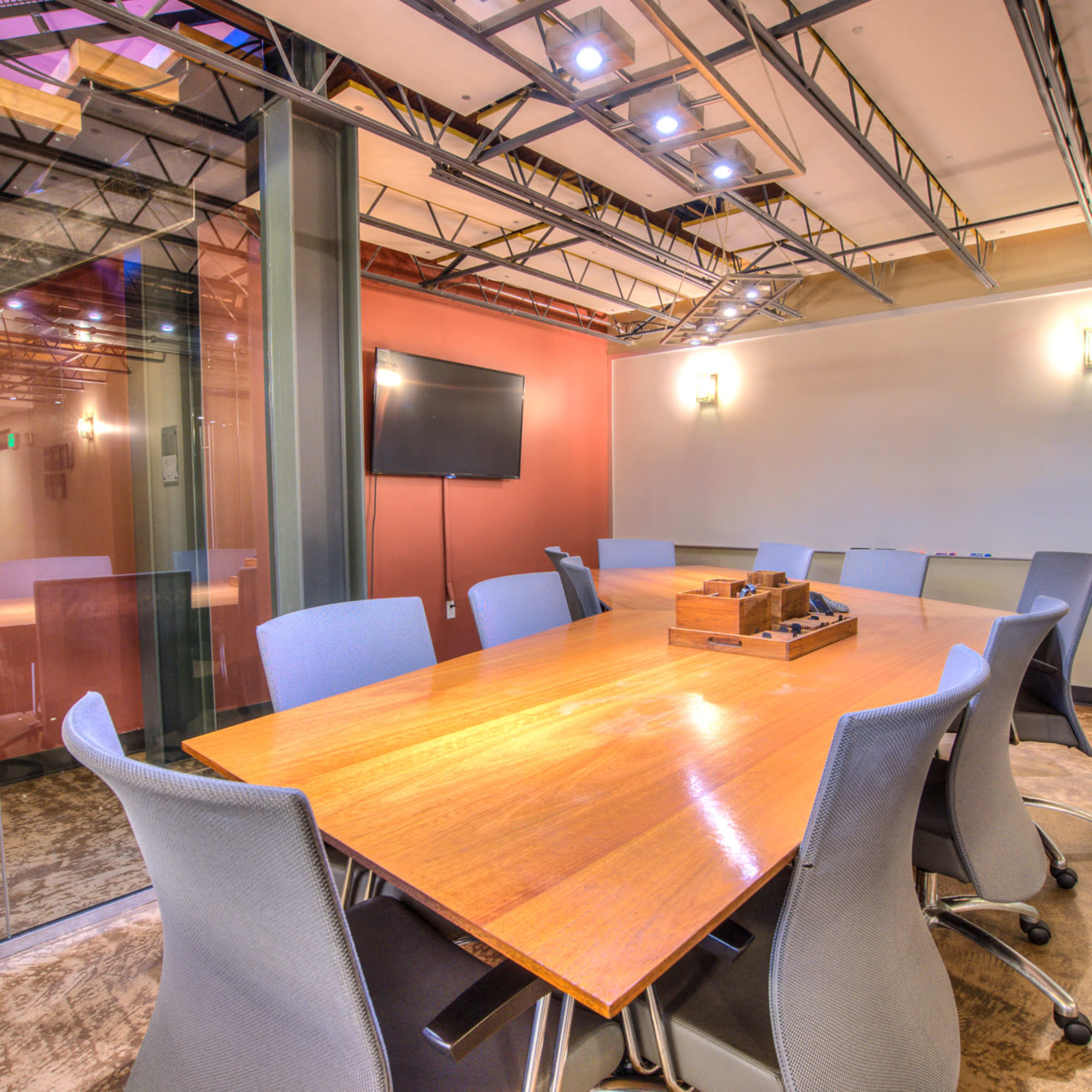 A conference room features a large wooden table surrounded by gray chairs, with a TV screen mounted on the wall and glass partitioning.