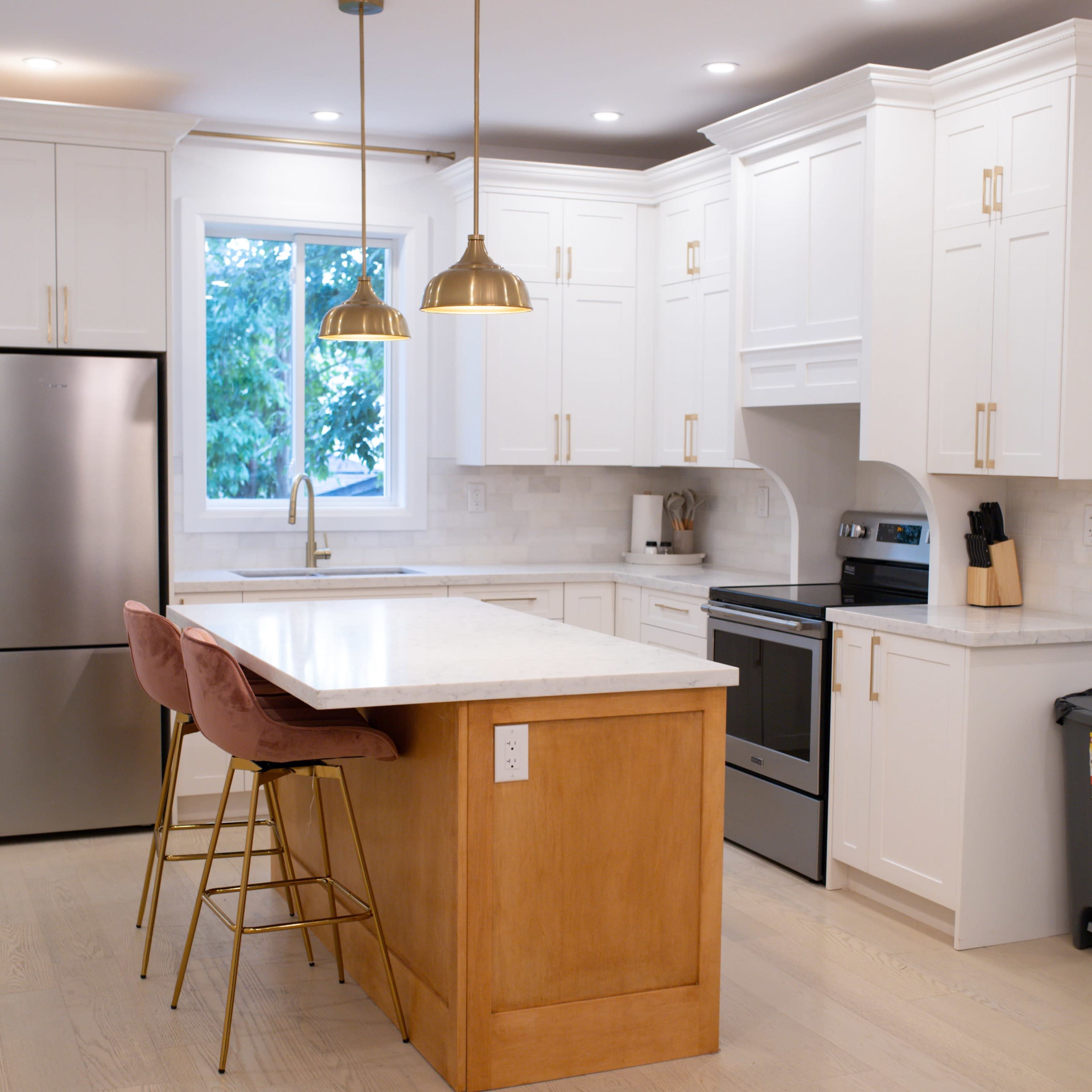 A modern kitchen features white cabinetry, stainless steel appliances, a large island with a wooden base, and two pink bar stools under gold pendant lights.