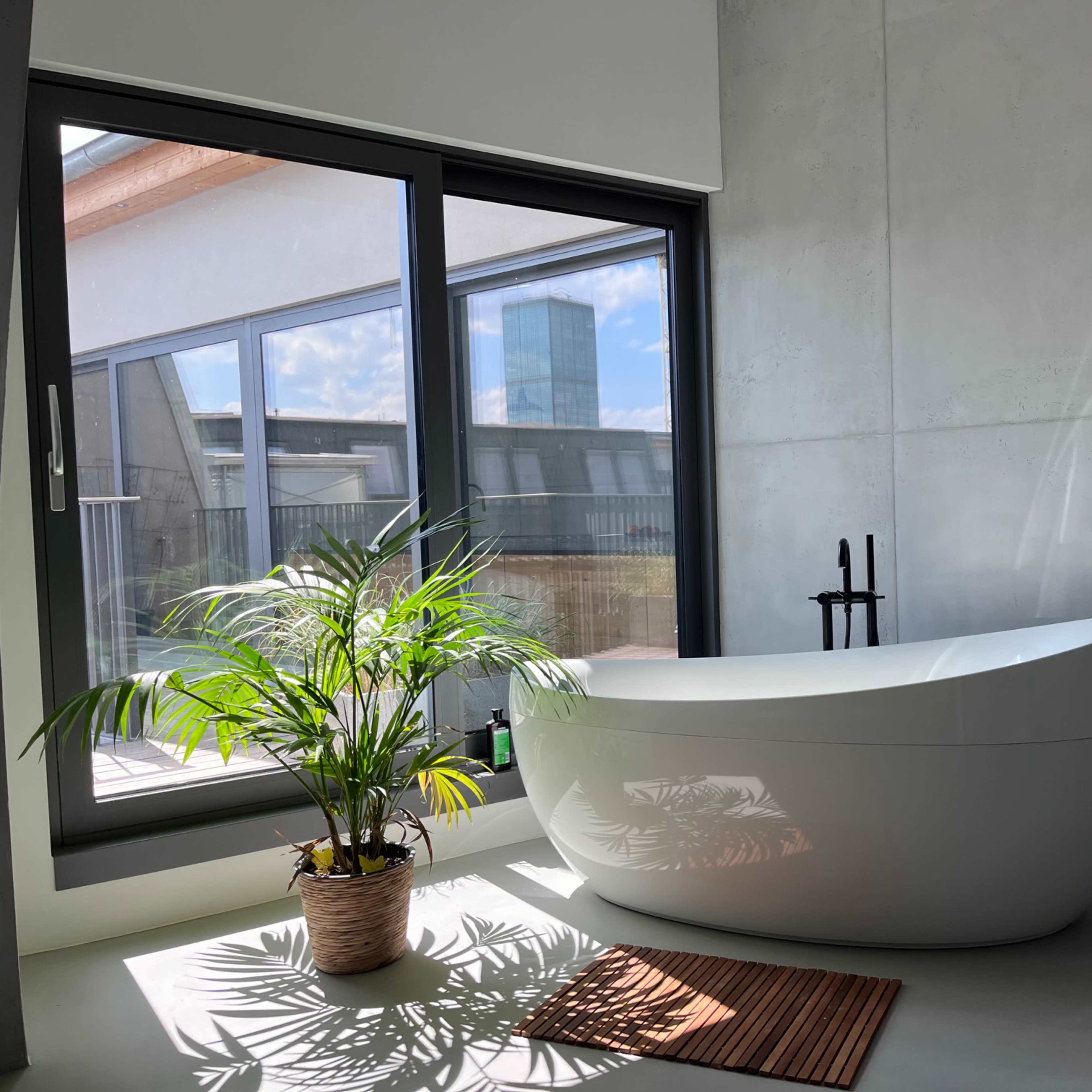 A modern bathroom featuring a freestanding bathtub beside a large window with a view, complemented by a potted plant and a bath mat on the floor.