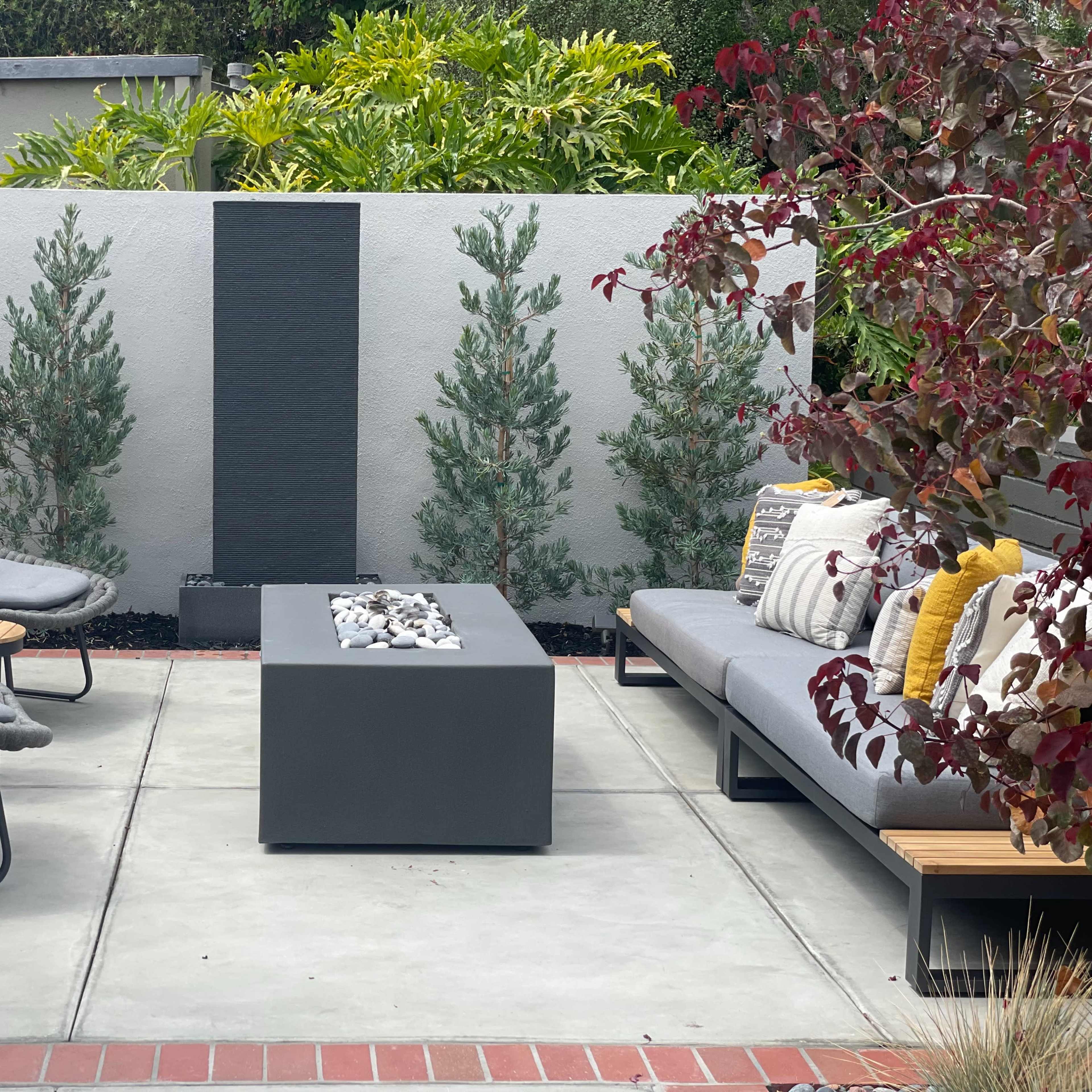 A modern outdoor seating area features two lounge chairs, a gray fire table, and a sofa, surrounded by landscaped greenery and a simple water feature.