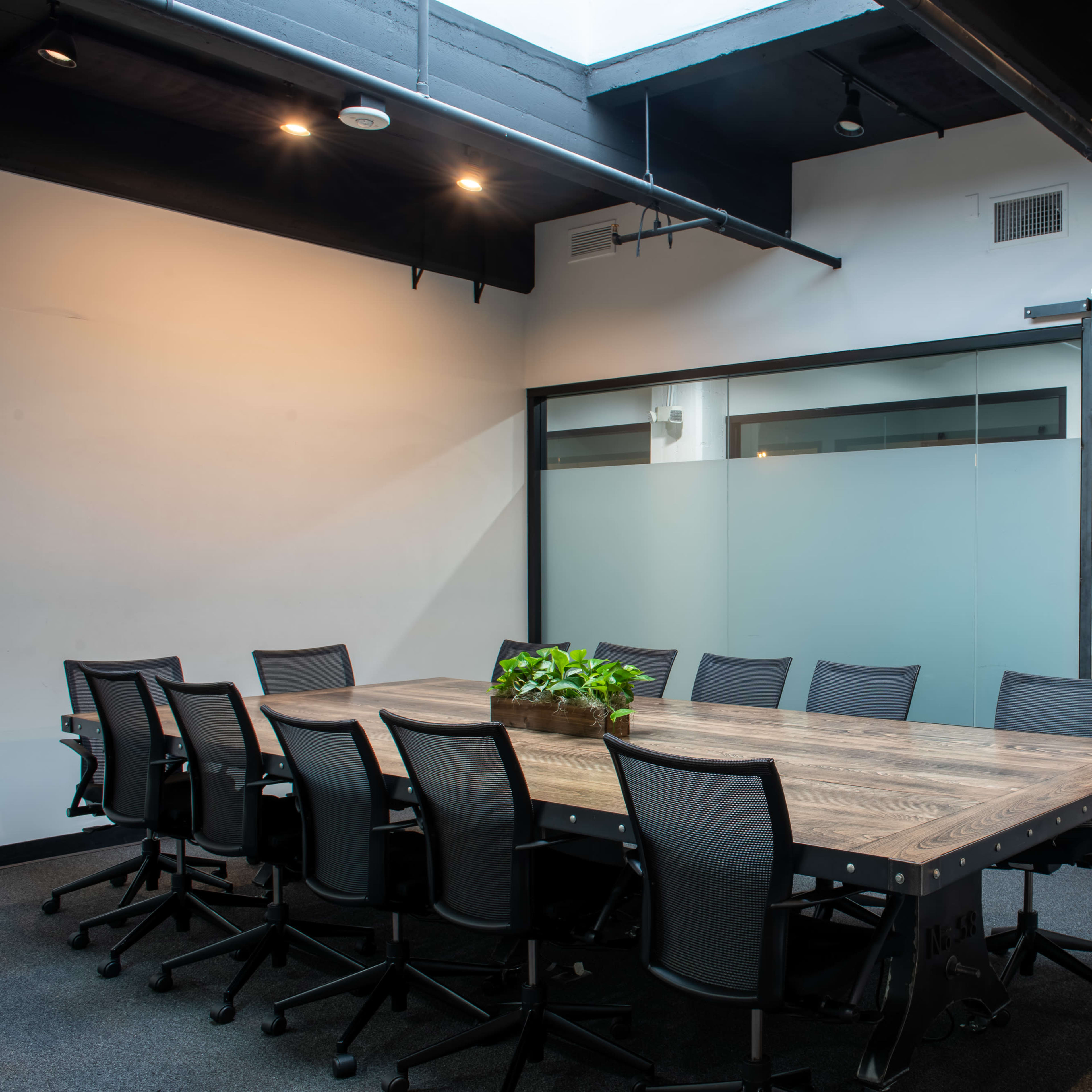 A conference room features a long wooden table surrounded by eight black chairs, with a sliding barn door and a plant centerpiece.