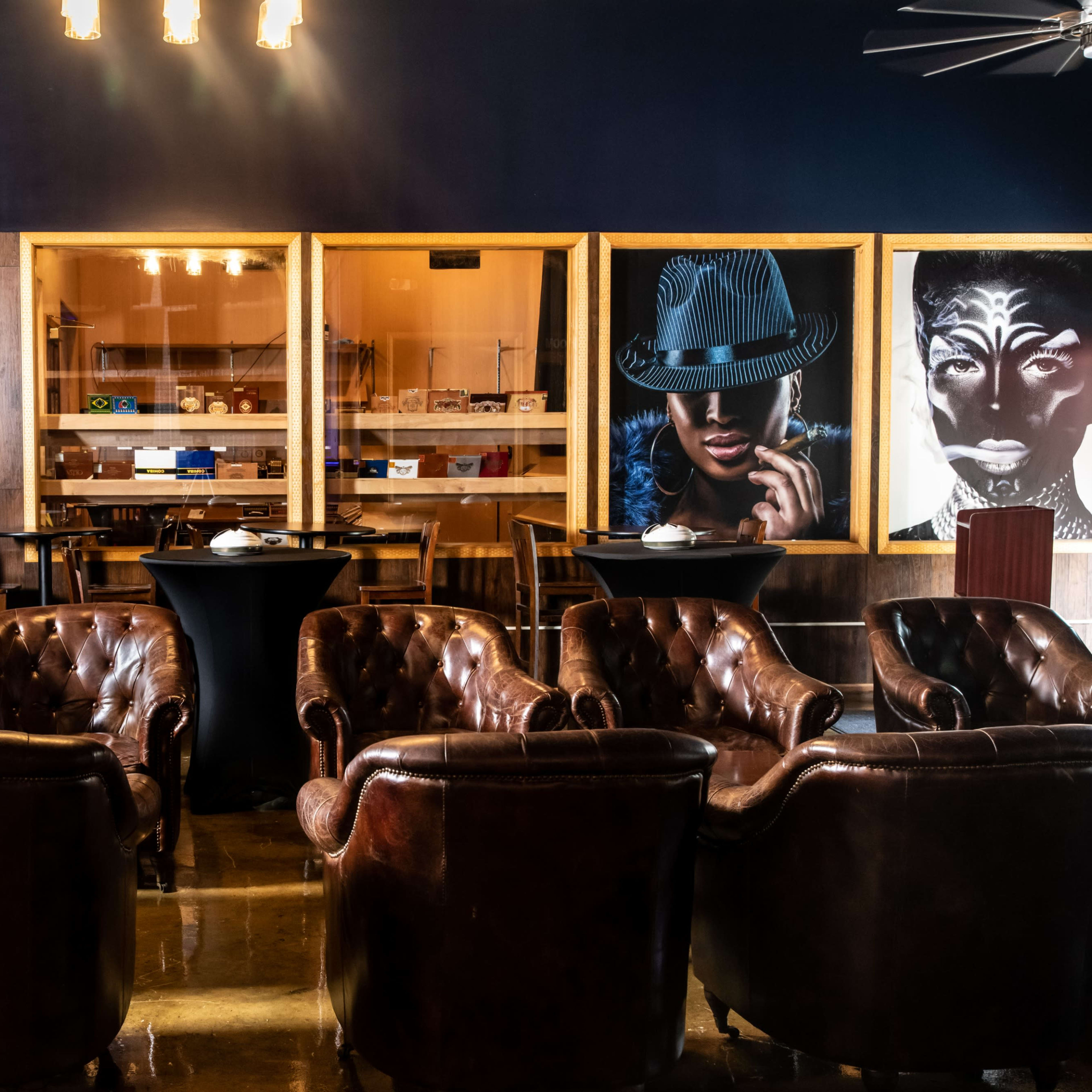 A lounge area with leather chairs arranged around high tables, showcasing artworks on the walls and a glass display in the background.