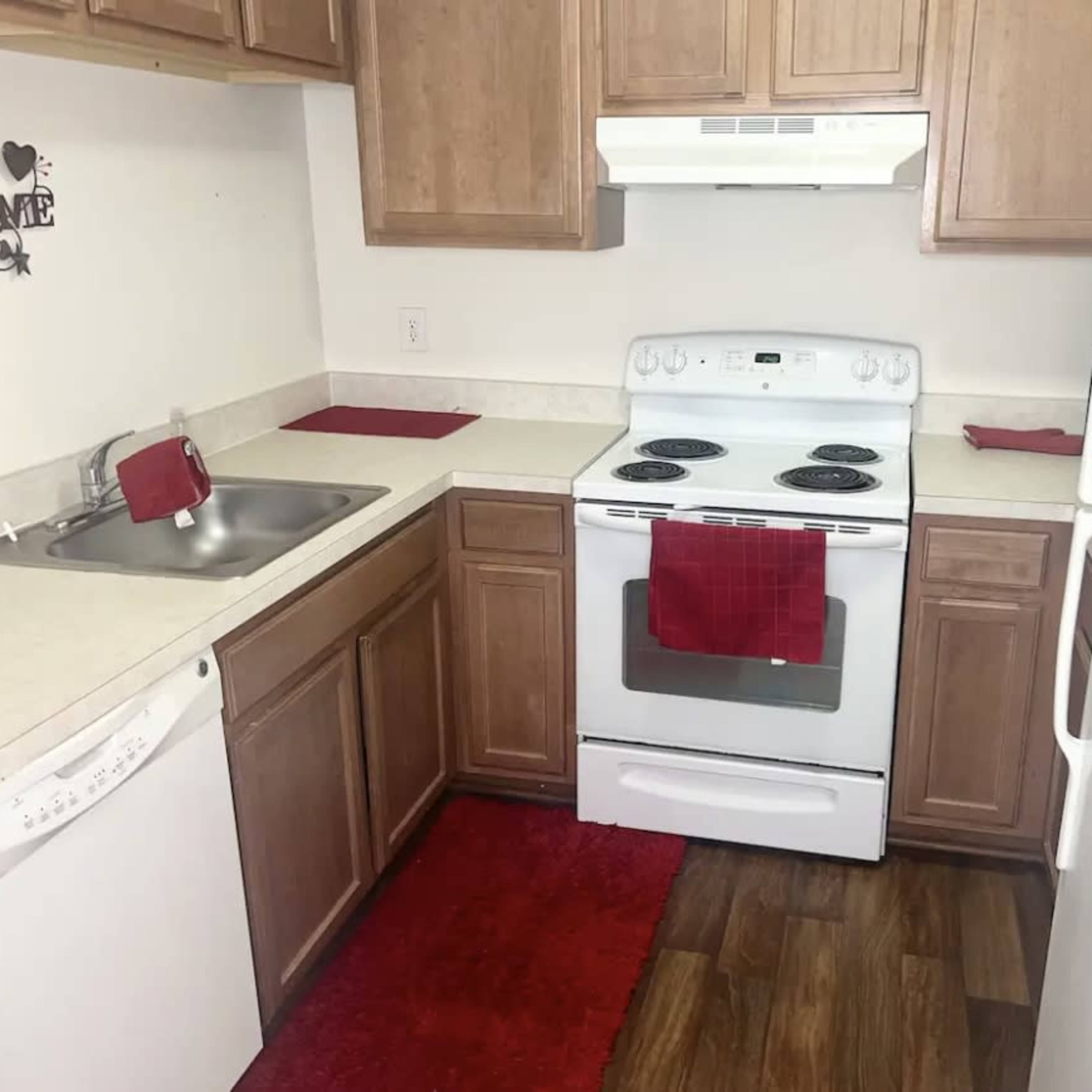 A kitchen with wooden cabinets, a white stove, a dishwasher, and a red dish towel on the counter, along with a matching red rug on the floor.