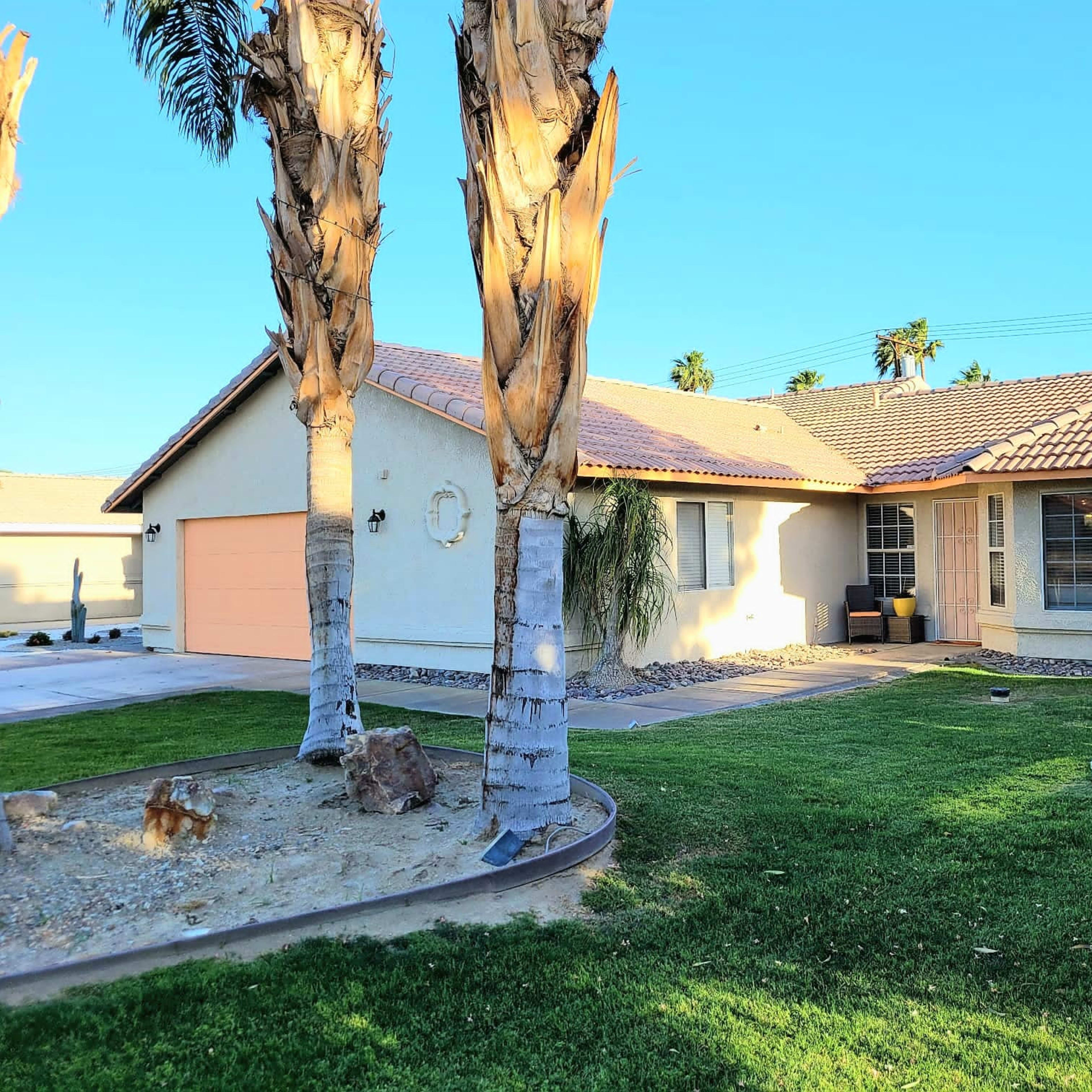 A single-story house with a beige exterior and a tiled roof sits in a yard flanked by palm trees.