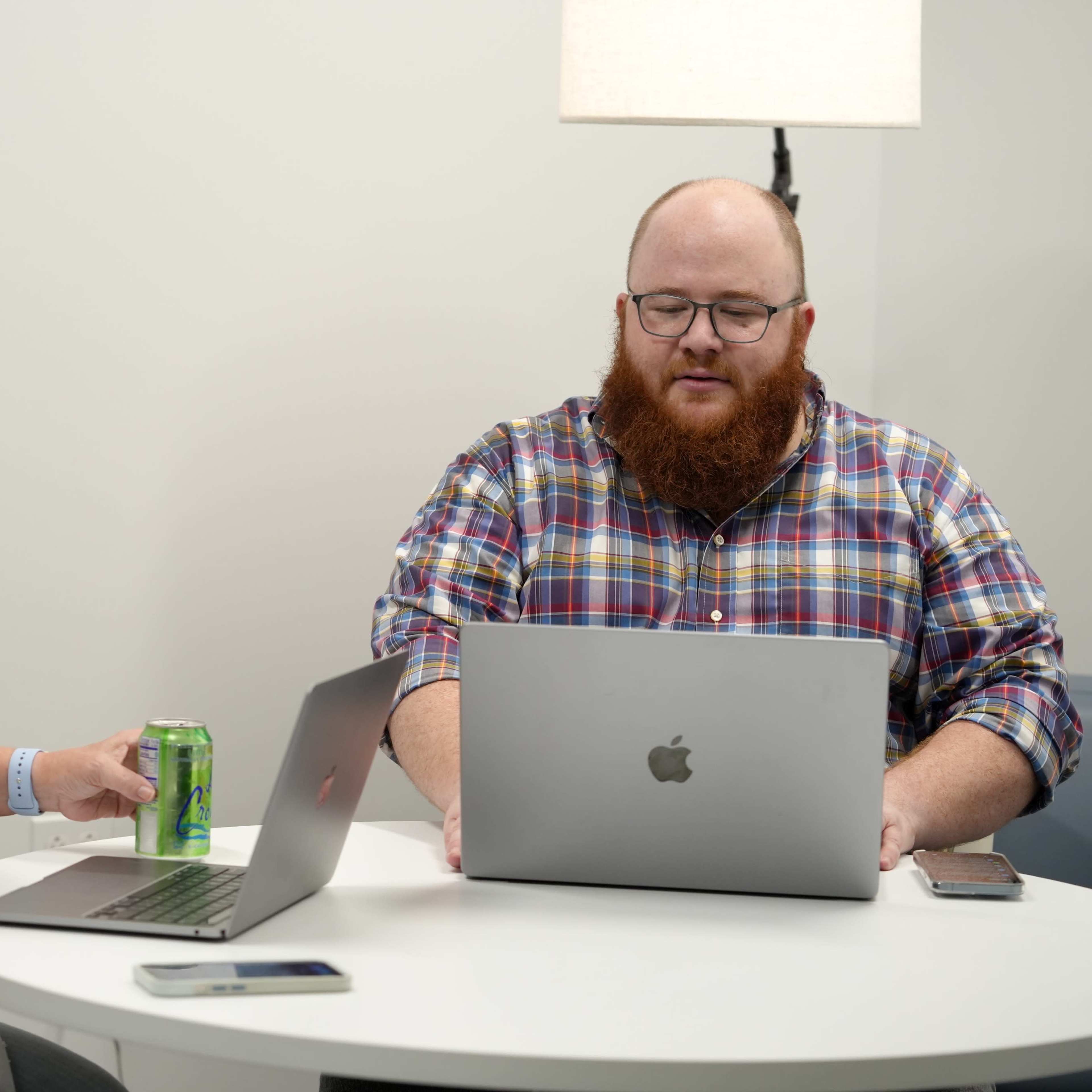 A woman sits at a table with a can of soda while a man in a plaid shirt works on a MacBook.