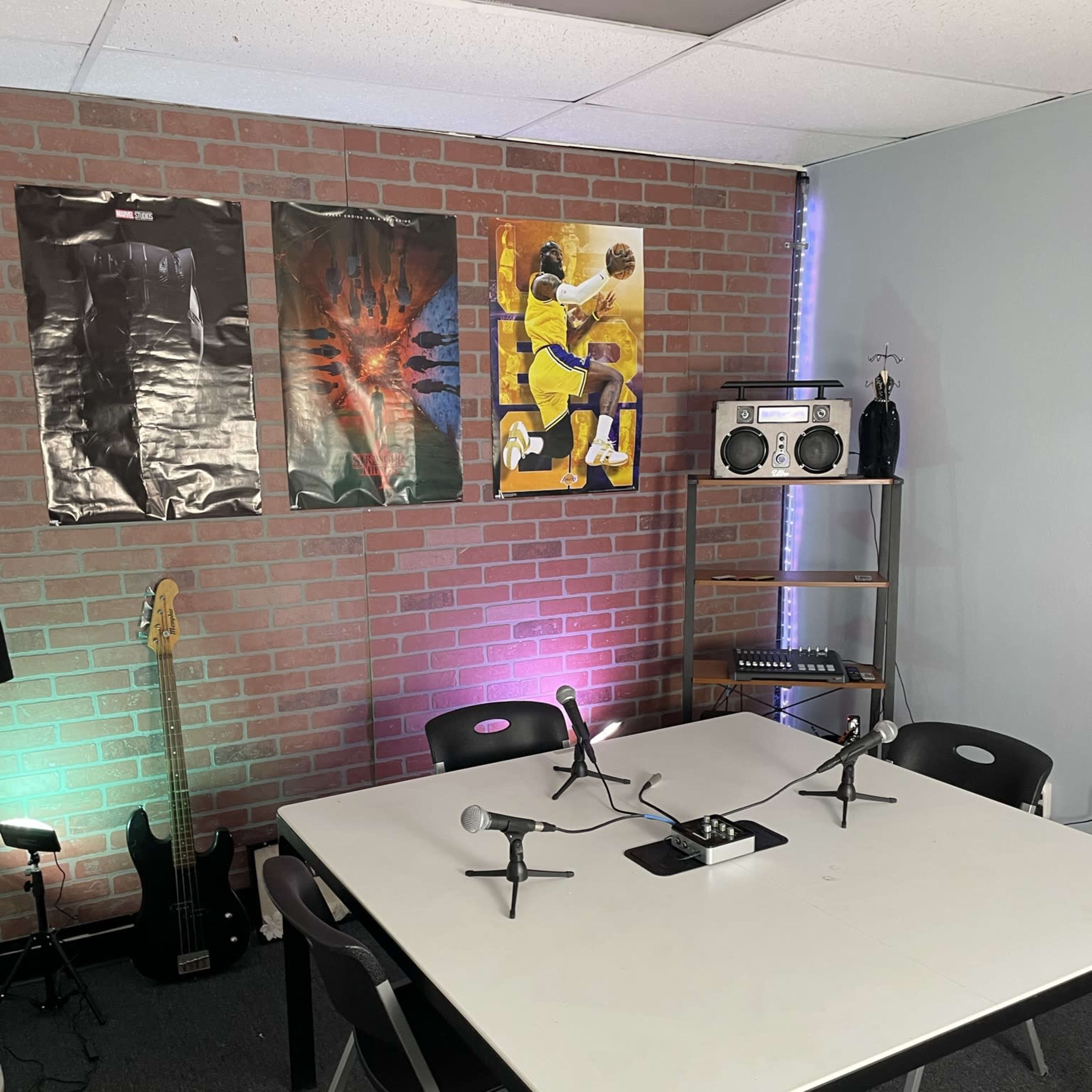 A small recording or meeting space with a table at the center, equipped with microphones, a boombox, and three posters on a brick wall, alongside a guitar and a shelf.