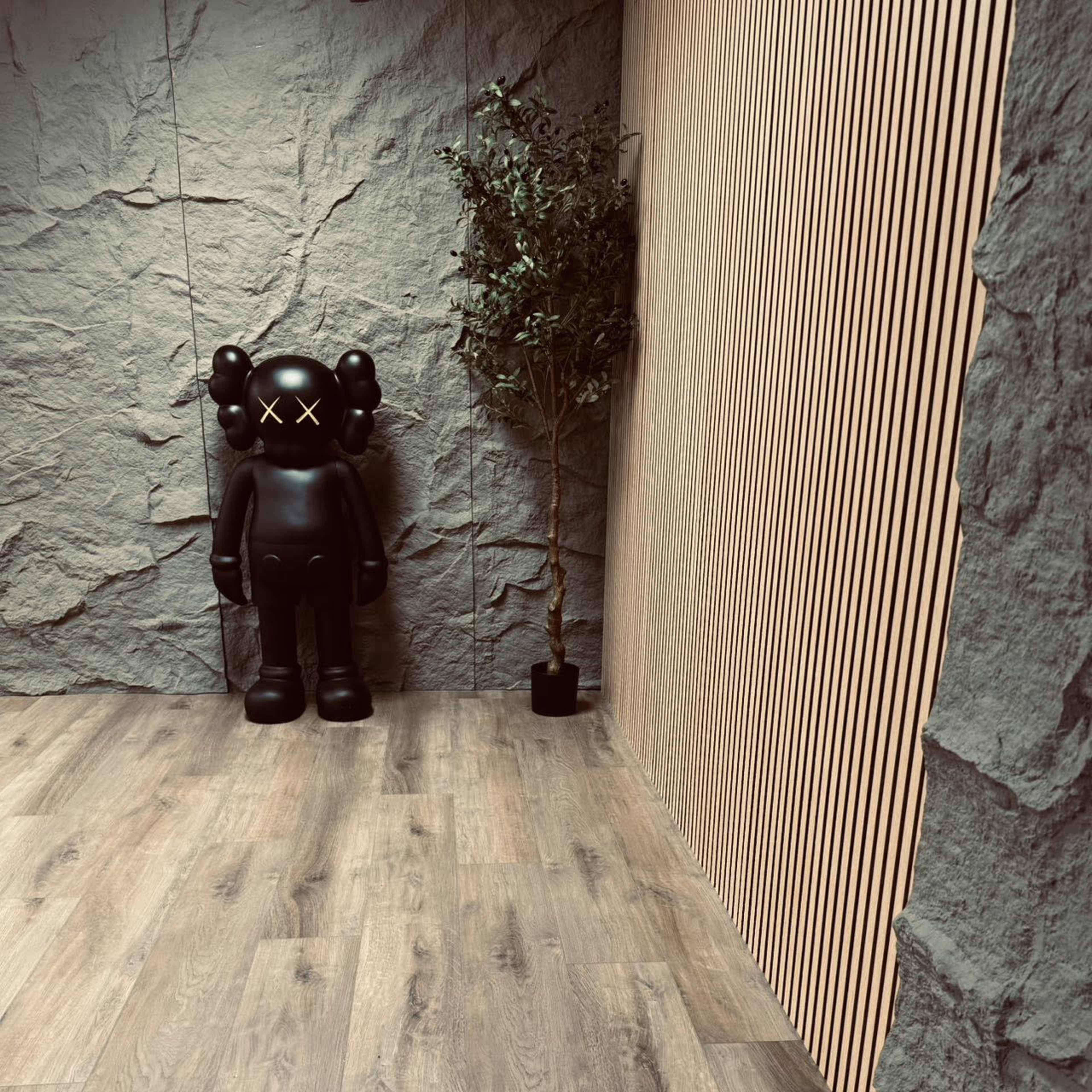 A black figurine stands against a textured stone wall next to a small potted plant in a minimalist indoor space featuring wooden flooring.