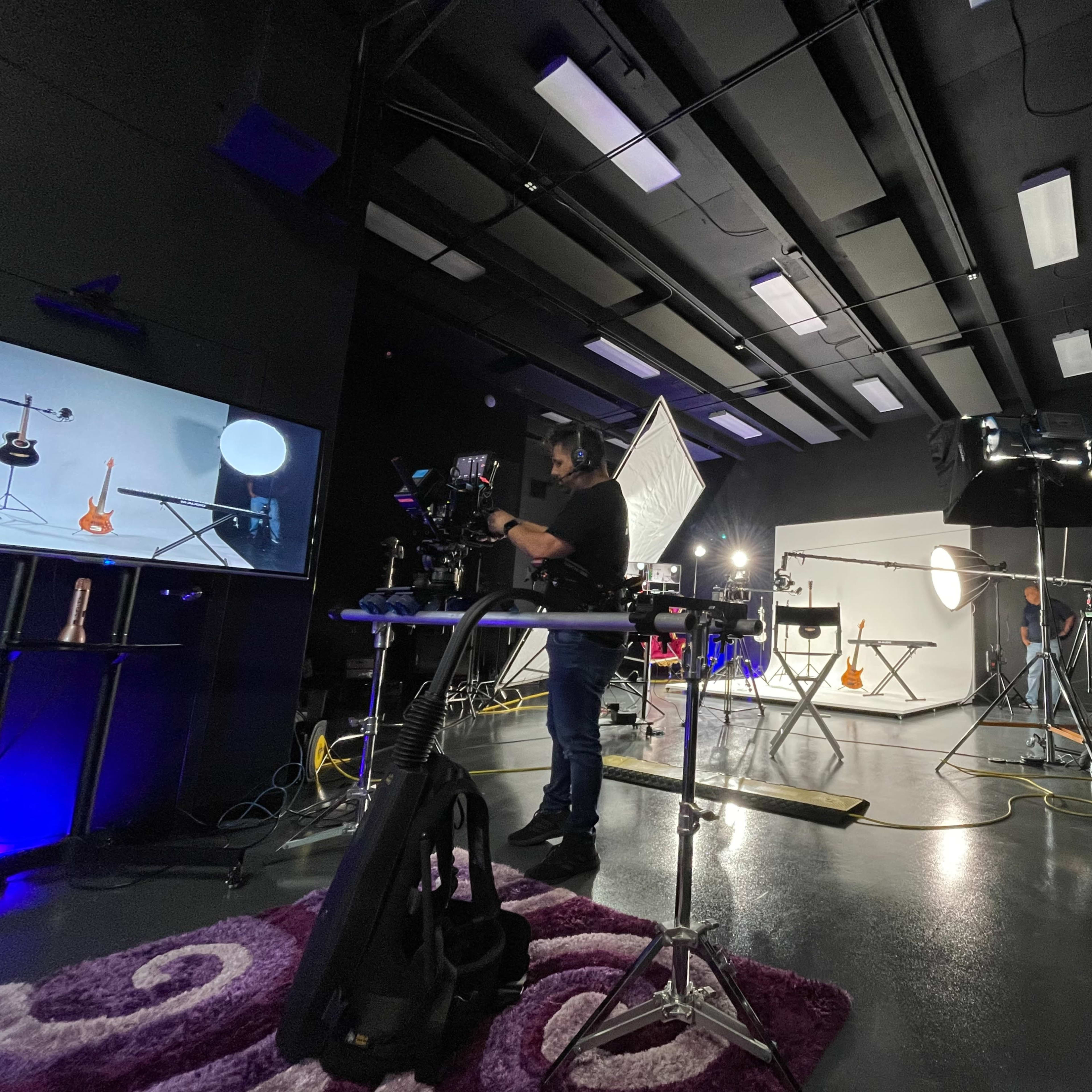 A photography studio is set up with various lighting equipment, a camera person preparing a shot, and a display screen showing a guitar setup.