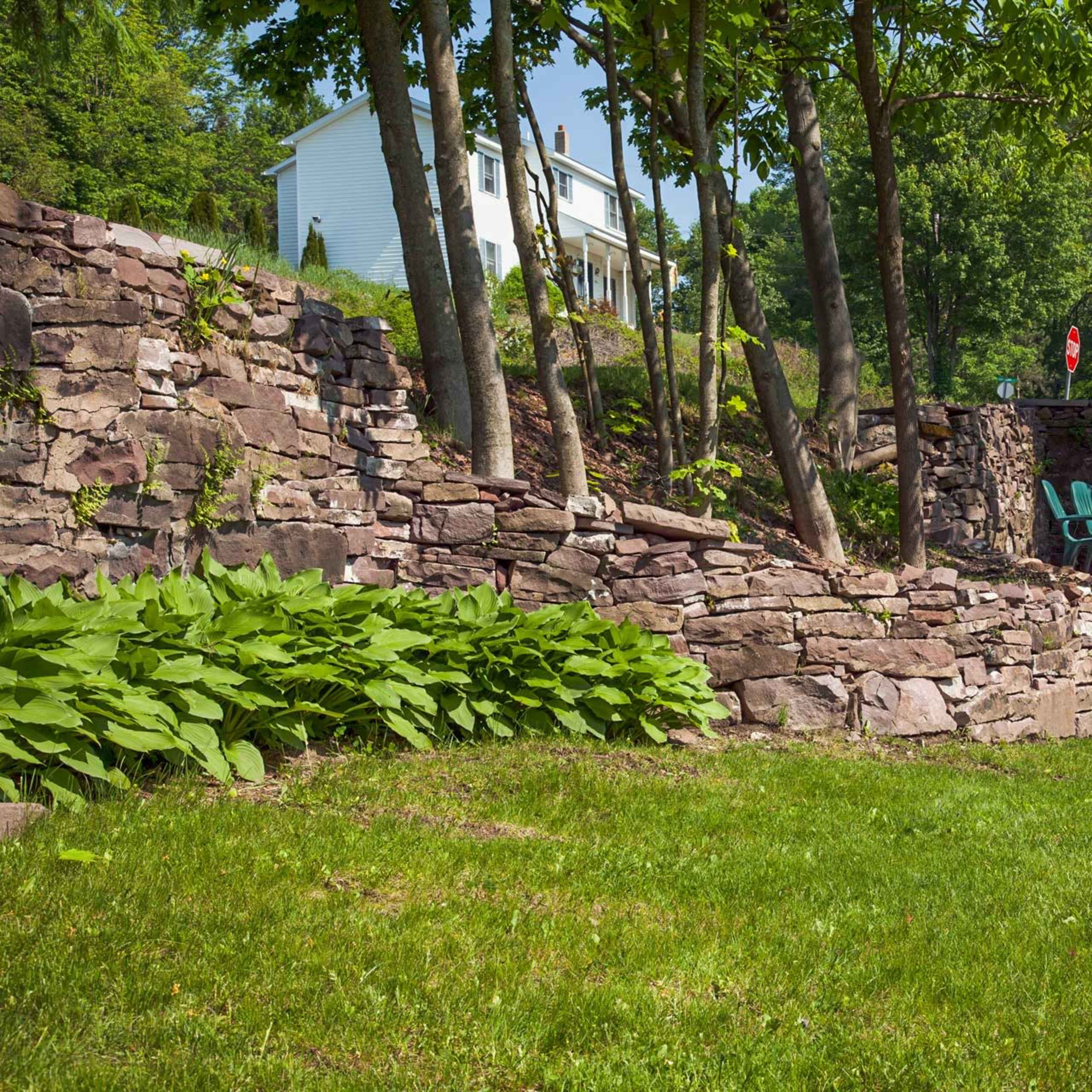 A landscaped yard with stone walls, green grass, and a pair of blue chairs under shaded trees.