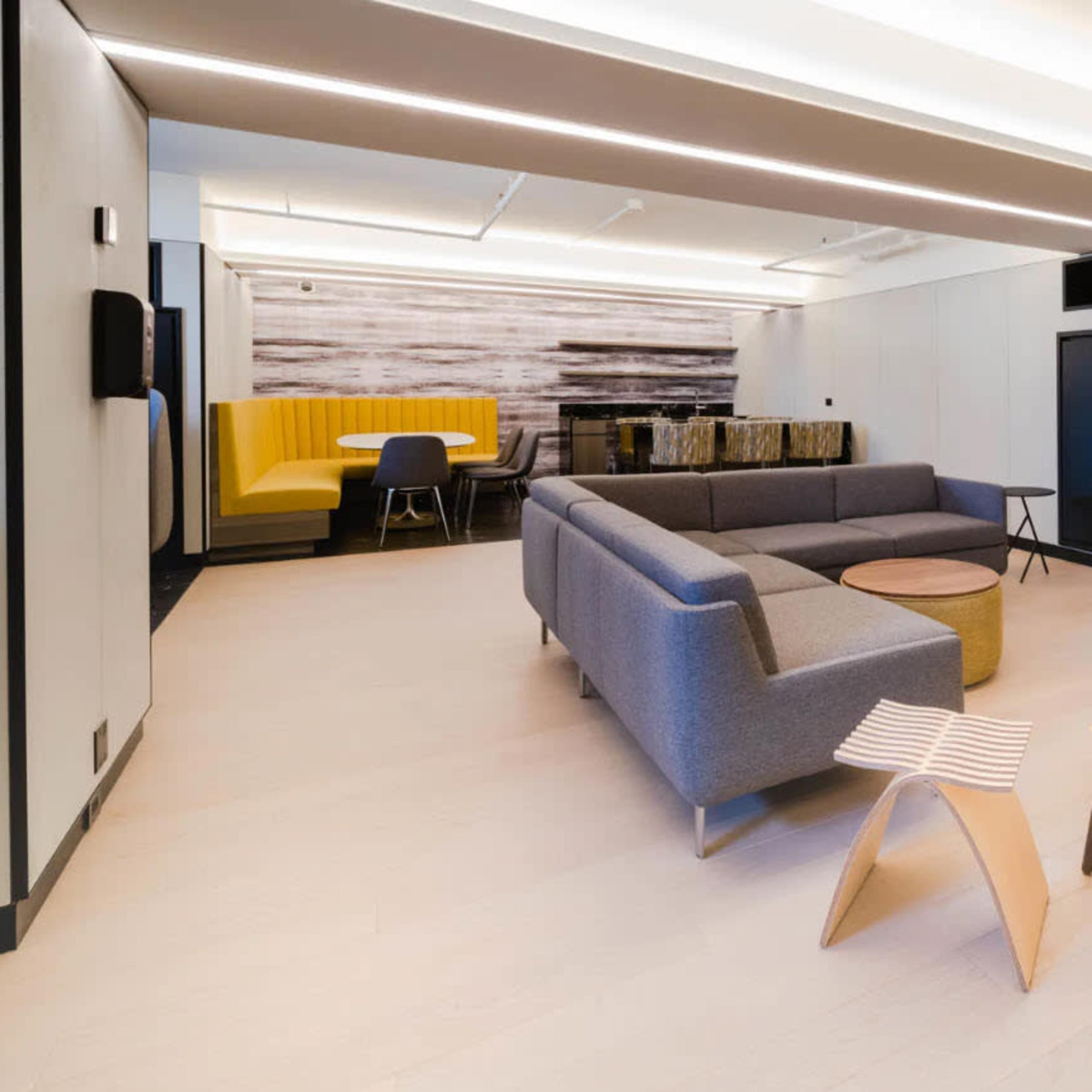 A modern, open lounge area featuring a gray sofa, yellow chairs, a round wooden coffee table, and a large television mounted on the wall.