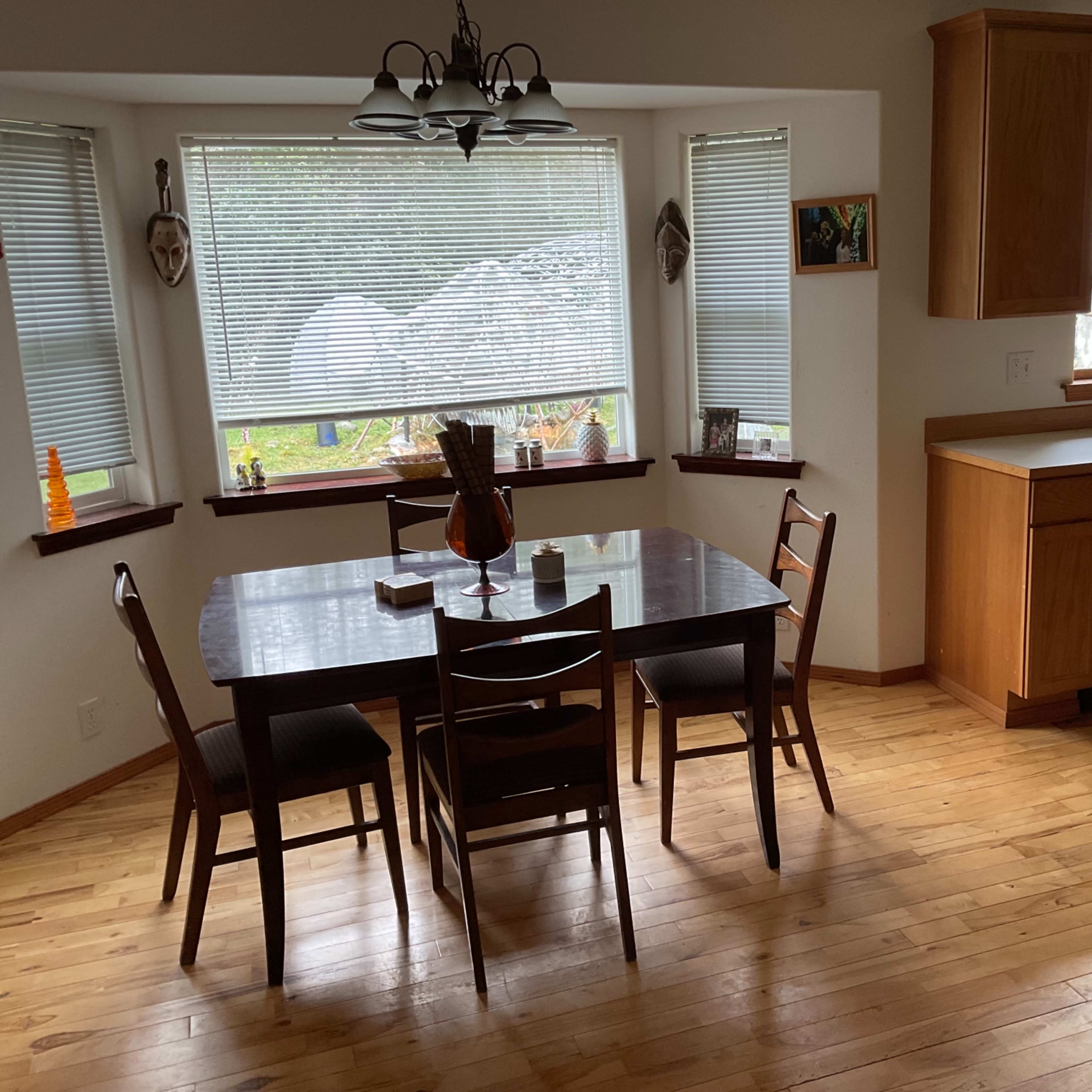 A wooden dining table with four chairs is positioned in a kitchen area featuring large windows and light-colored cabinetry.