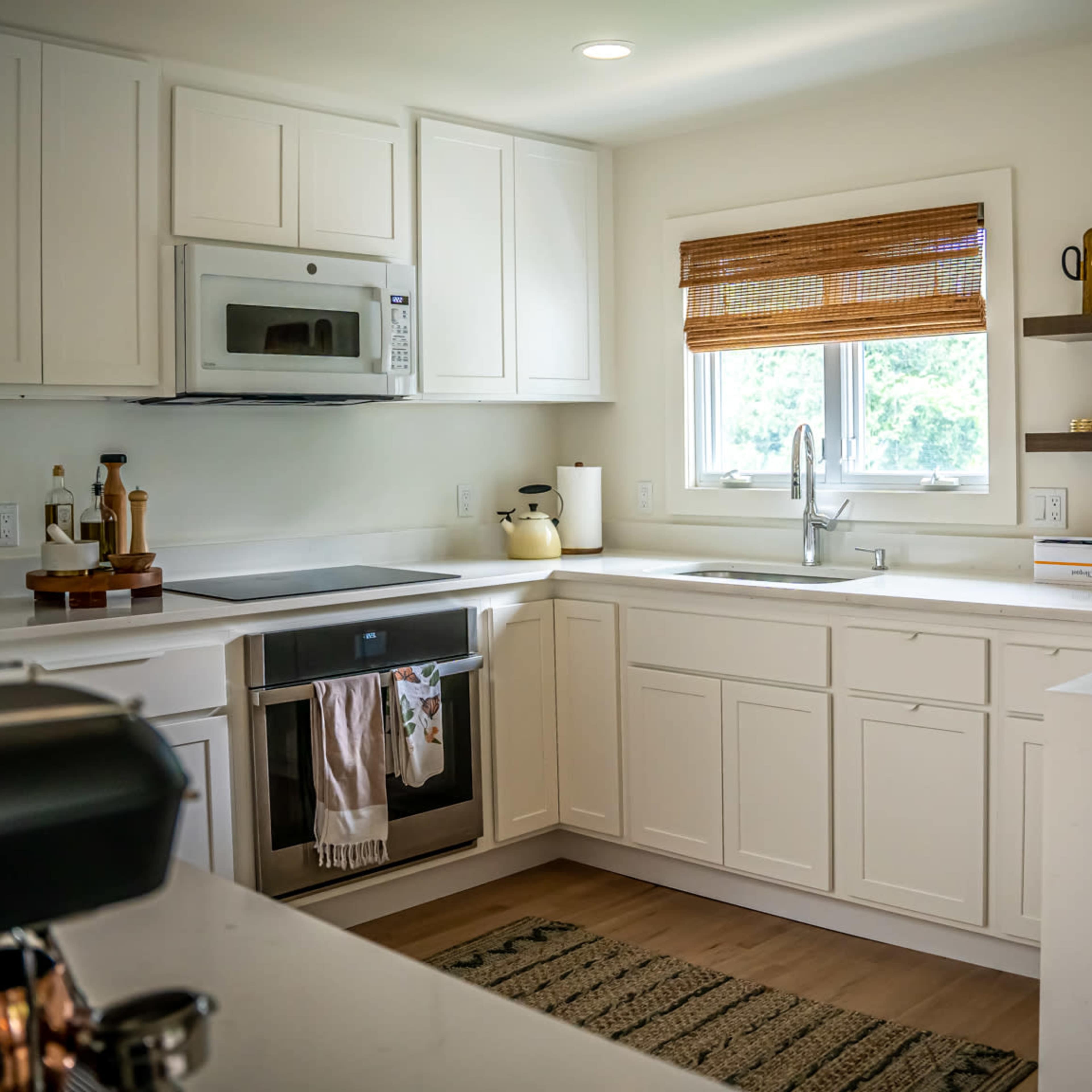 A bright kitchen features white cabinetry, a modern stove, a stainless steel microwave, and a window with wooden blinds.