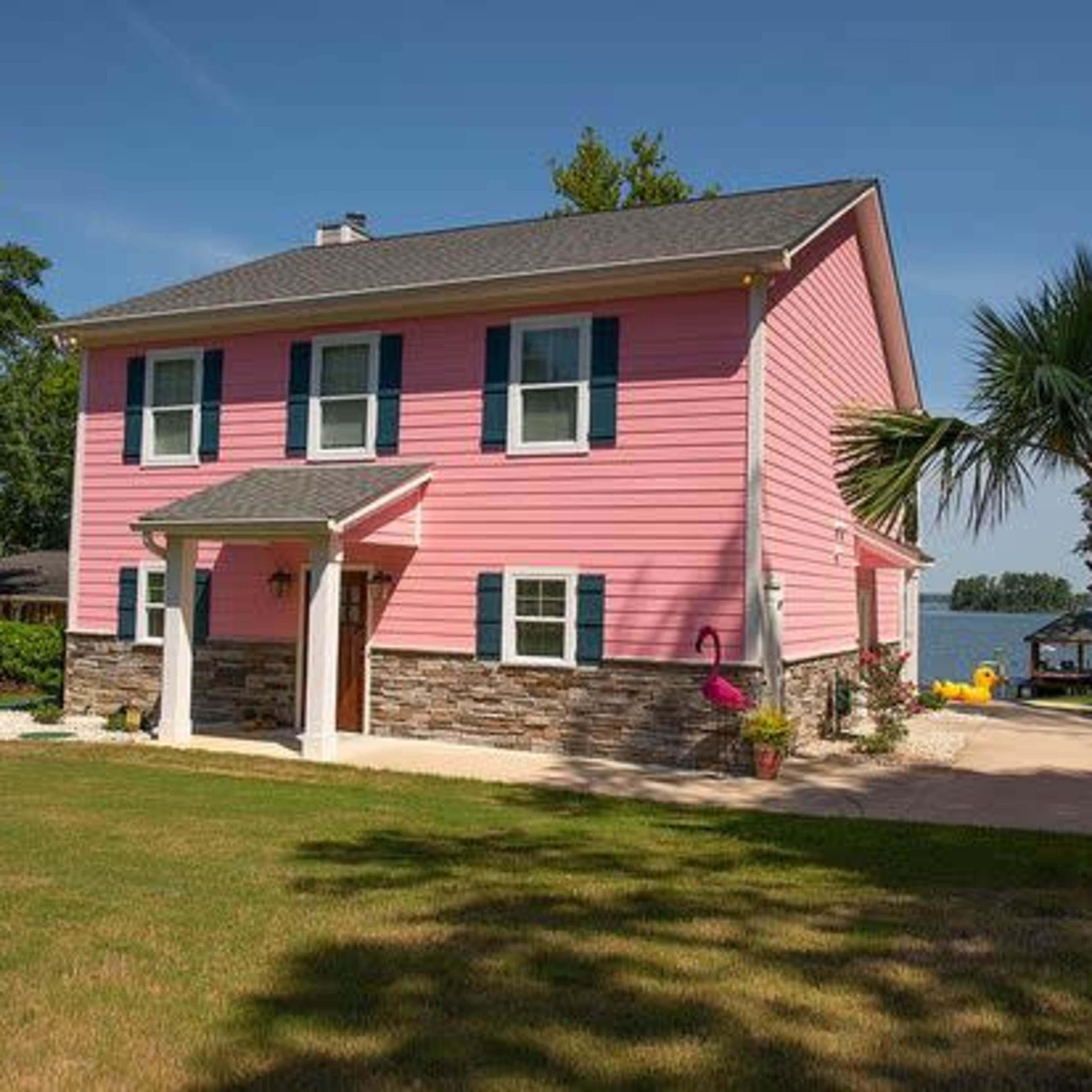 A pink two-story house with stone accents and green shutters is situated near a lake, featuring a well-maintained lawn and palm trees.
