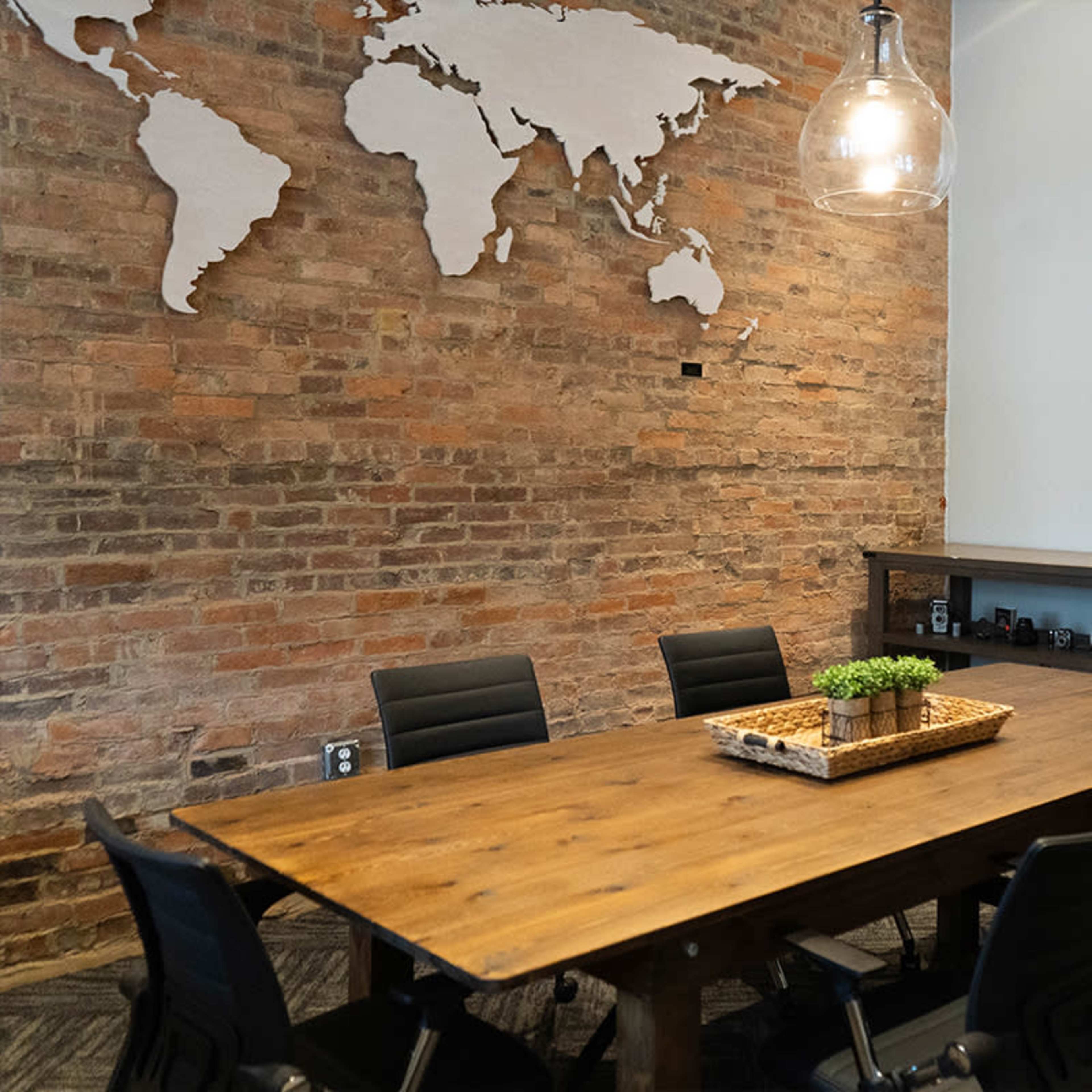A conference room with a large wooden table surrounded by black chairs, a world map on the brick wall, and a television mounted on the adjacent wall.